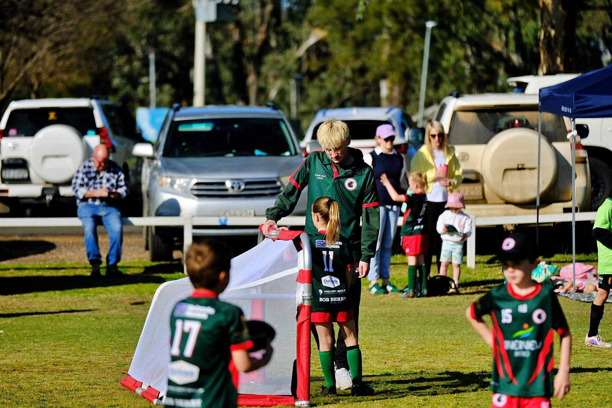 Children playing a soccer game outdoors with spectators and cars in the background on a sunny day.