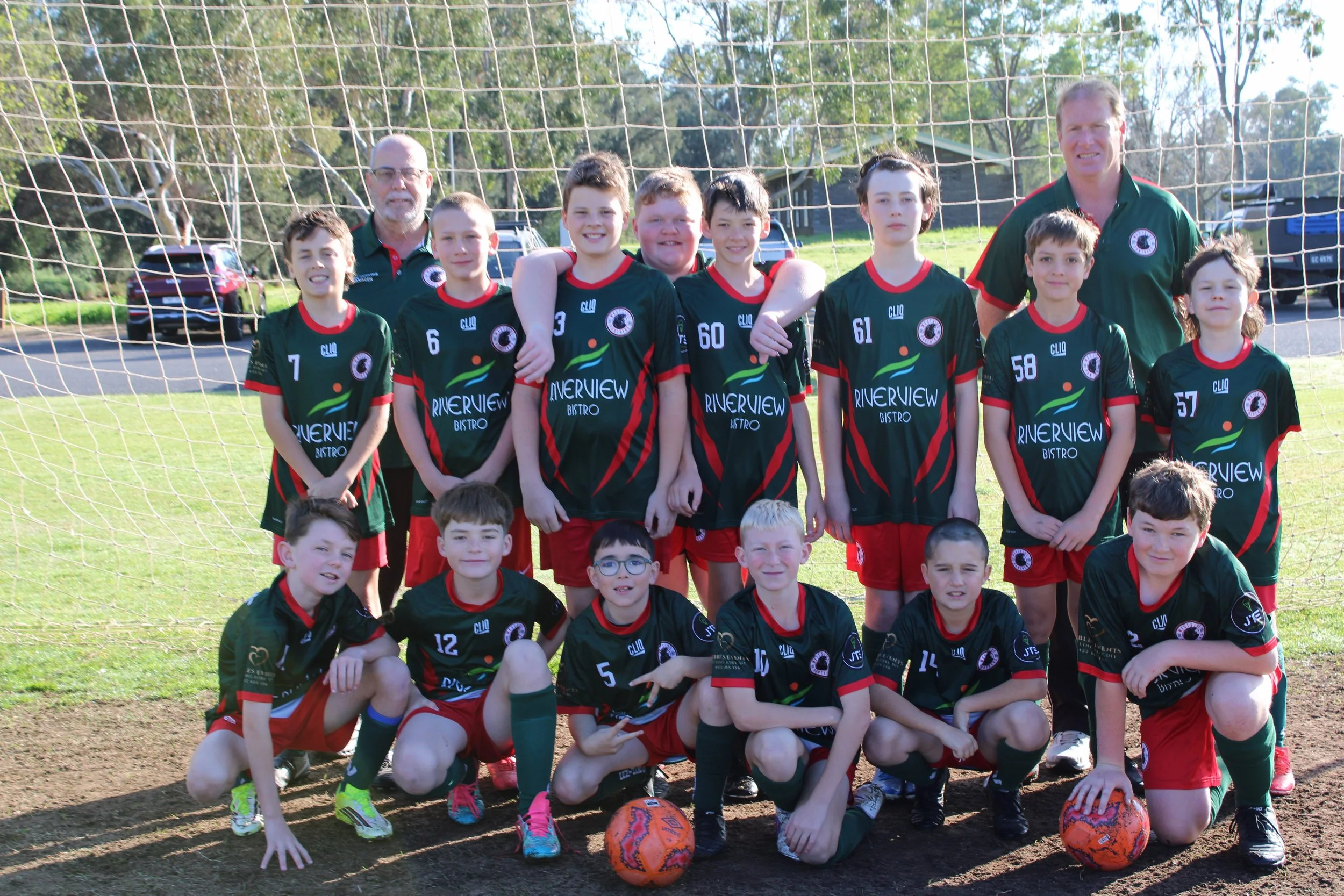 A youth soccer team of ten players and two coaches posing on a soccer field, with a goalpost behind them, outdoor setting, and several vehicles parked nearby.