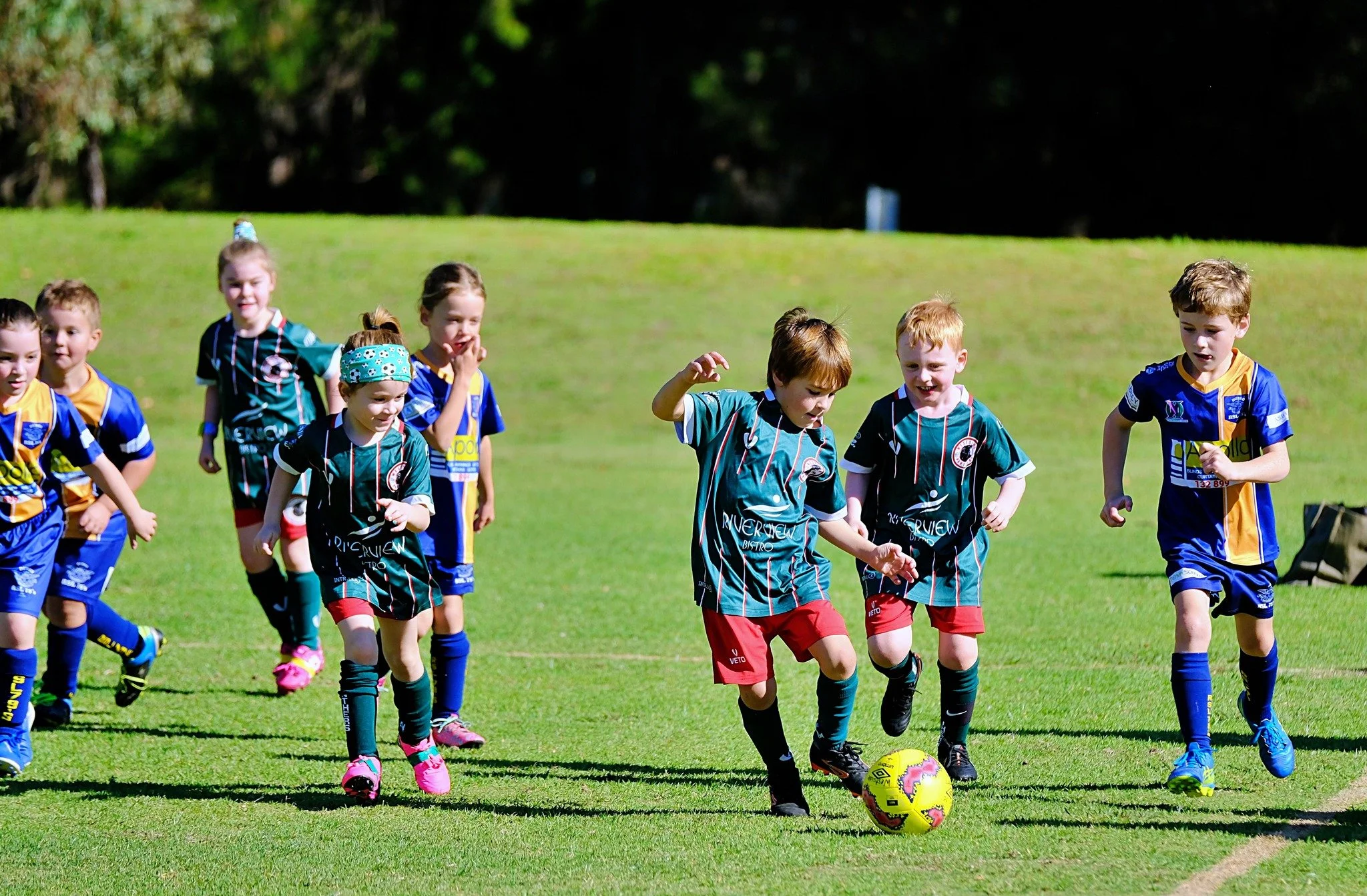 Children playing soccer on a green field, some running and one kicking a yellow soccer ball, with trees in the background.