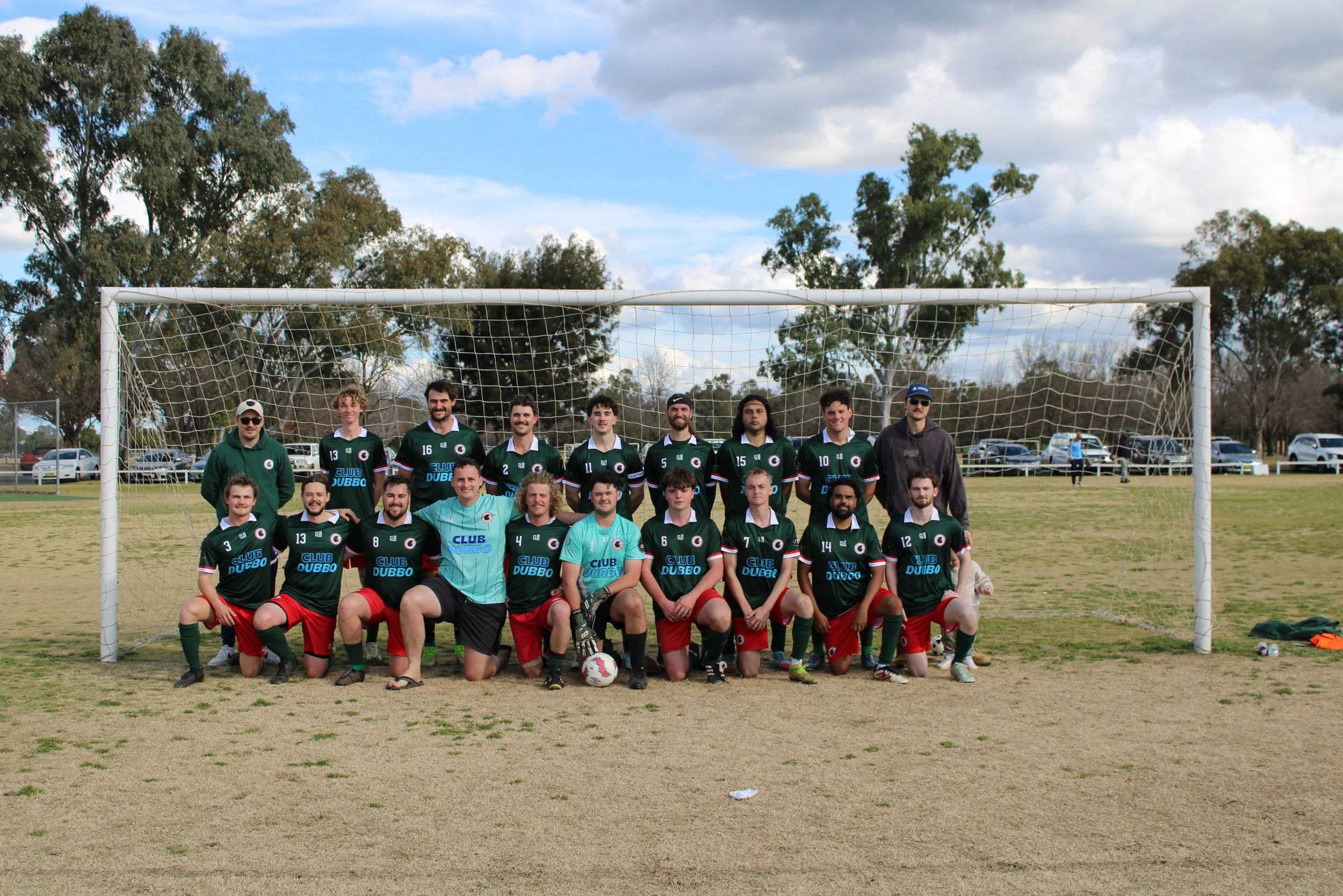 A group of soccer players and coaches posing in front of a goal on a grassy field during daytime. There are cars parked in the background, and trees and a partly cloudy sky are visible.