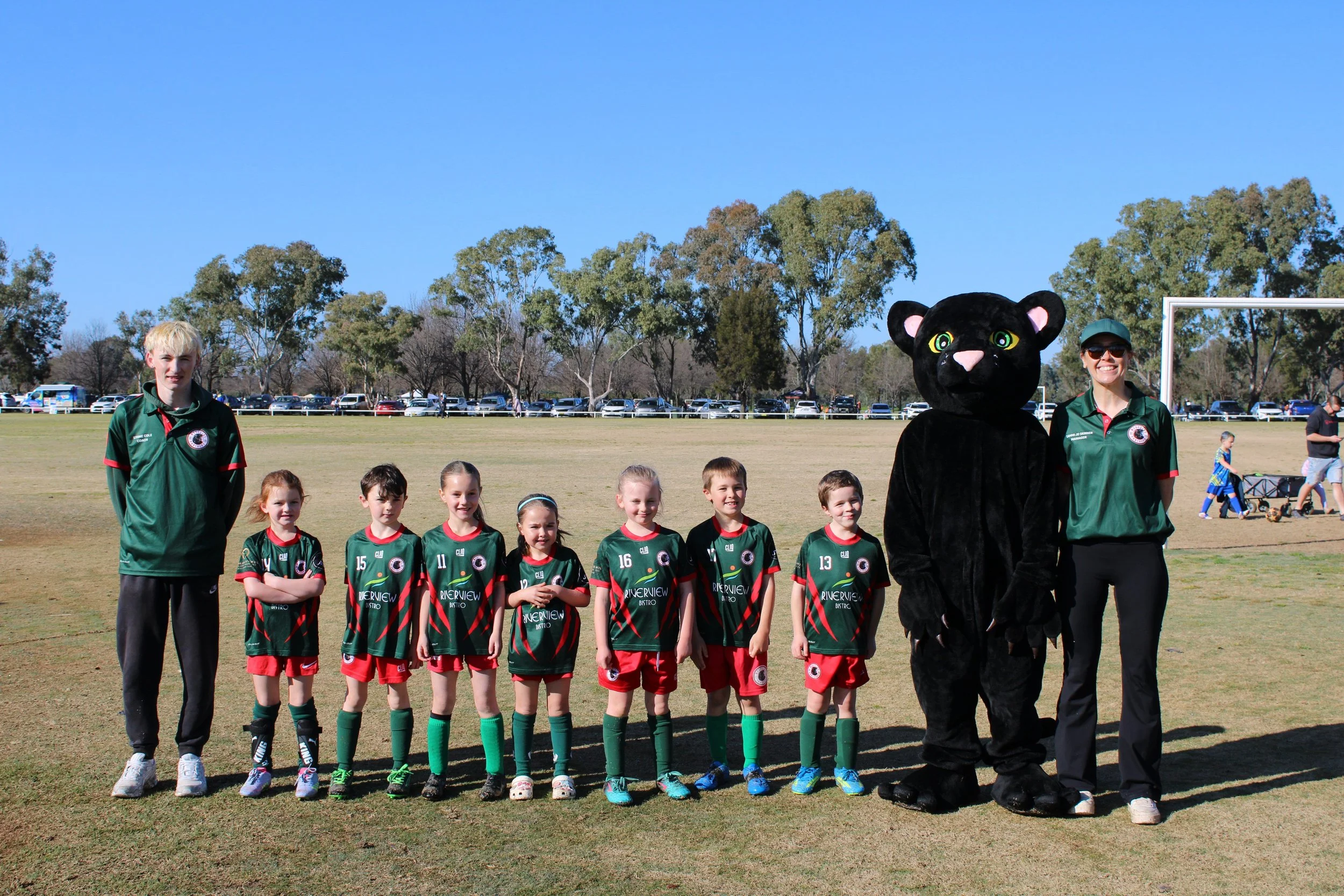 Children in soccer uniforms standing in a line on a sports field with a mascot in a black panther costume and two adults in green shirts, under a clear blue sky.