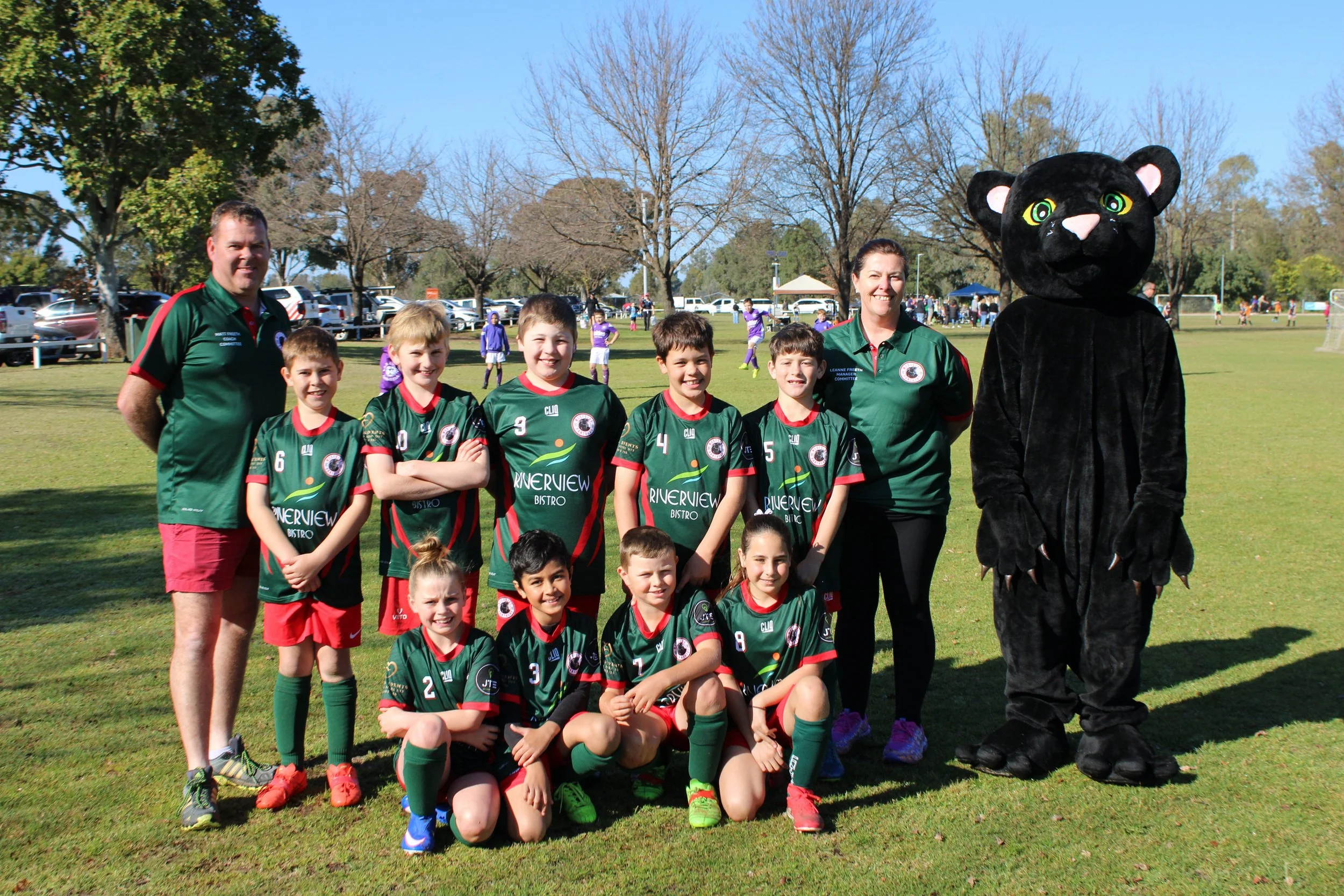 A group of young soccer players and their coaches, including a person in a black panther mascot costume, posing outdoors on a soccer field with trees and other players in the background.