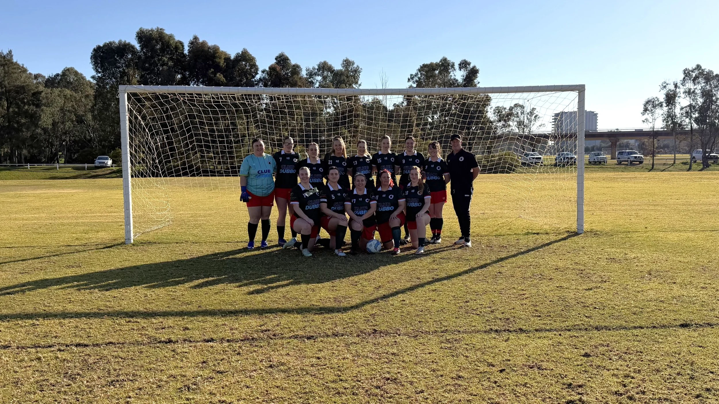 A women's soccer team posing for a group photo in front of goal on a grassy field during daytime. The team members are wearing black jerseys with blue and white accents, red shorts, and socks, with some kneeling in front and others standing behind. A person on the right is wearing black sportswear, and the goalkeeper on the left is dressed in a blue jersey and gloves. There are trees and parked cars in the background.