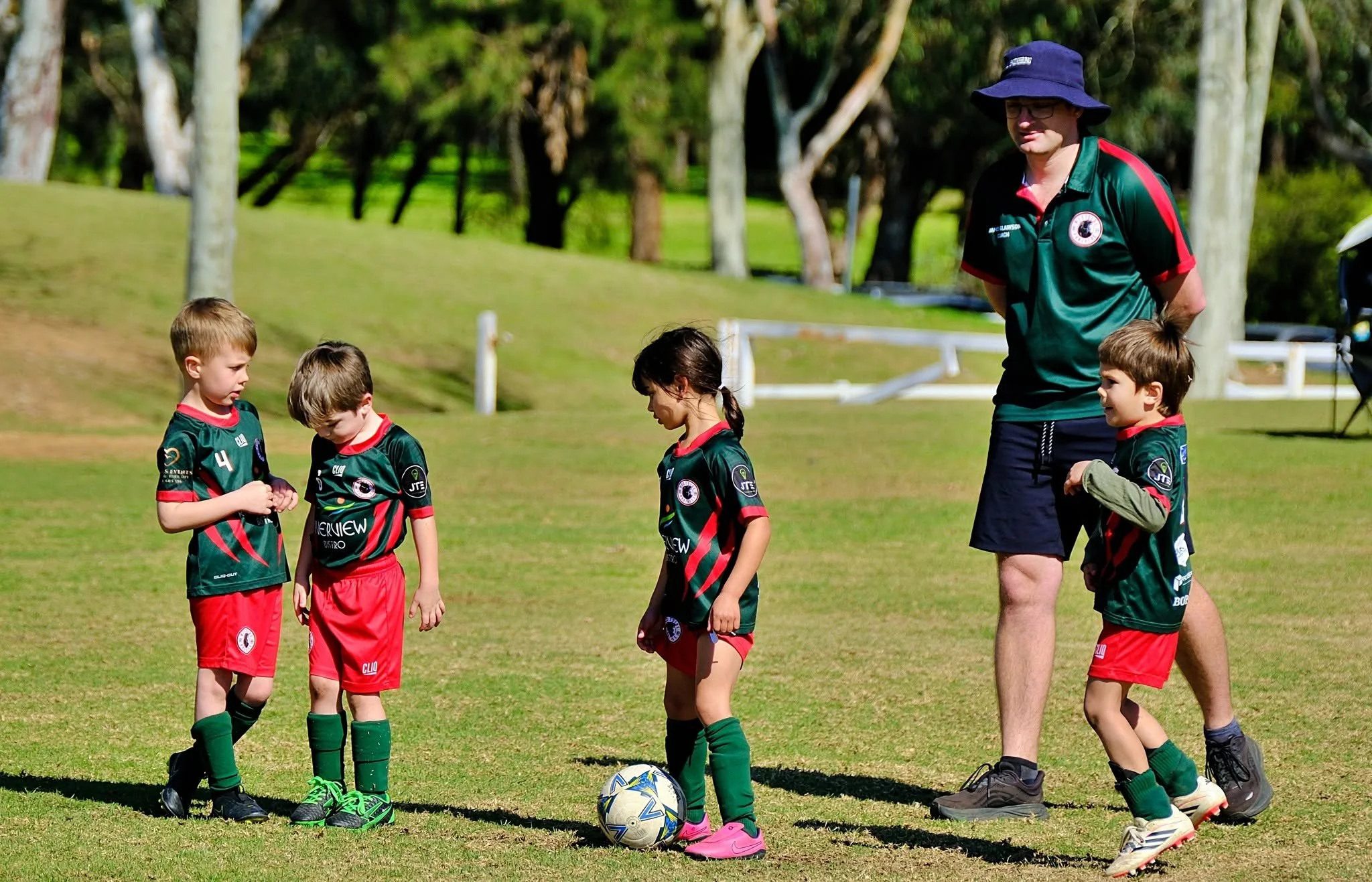 Children in a soccer team standing on a grassy field with a coach, preparing for a game and soccer balls visible.