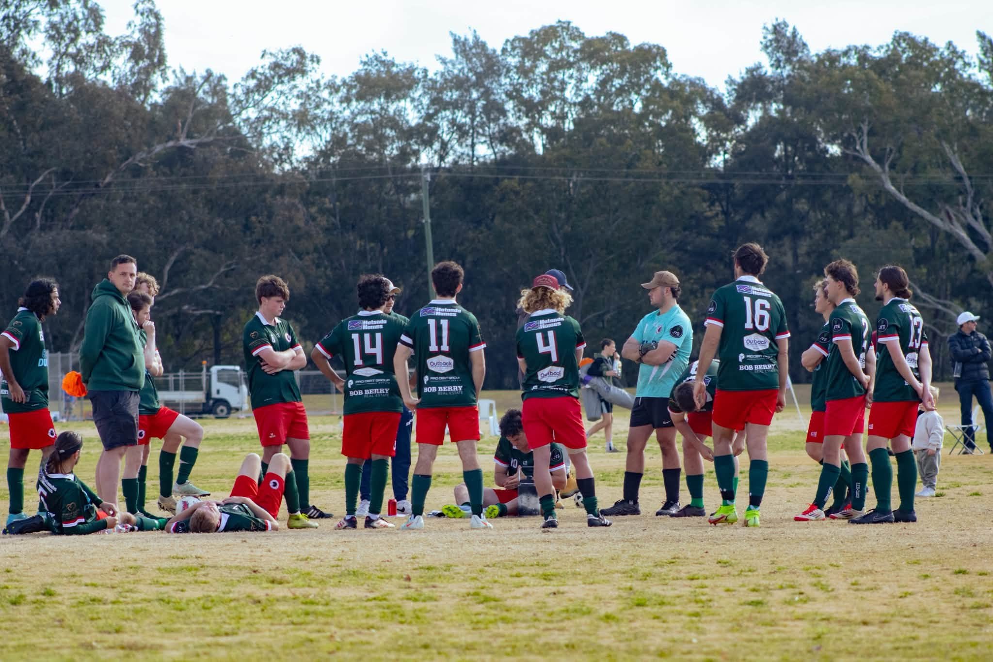 A group of rugby players gathered on a field, some standing and some sitting, with a coach or team staff member nearby, during a game or practice session with trees in the background.