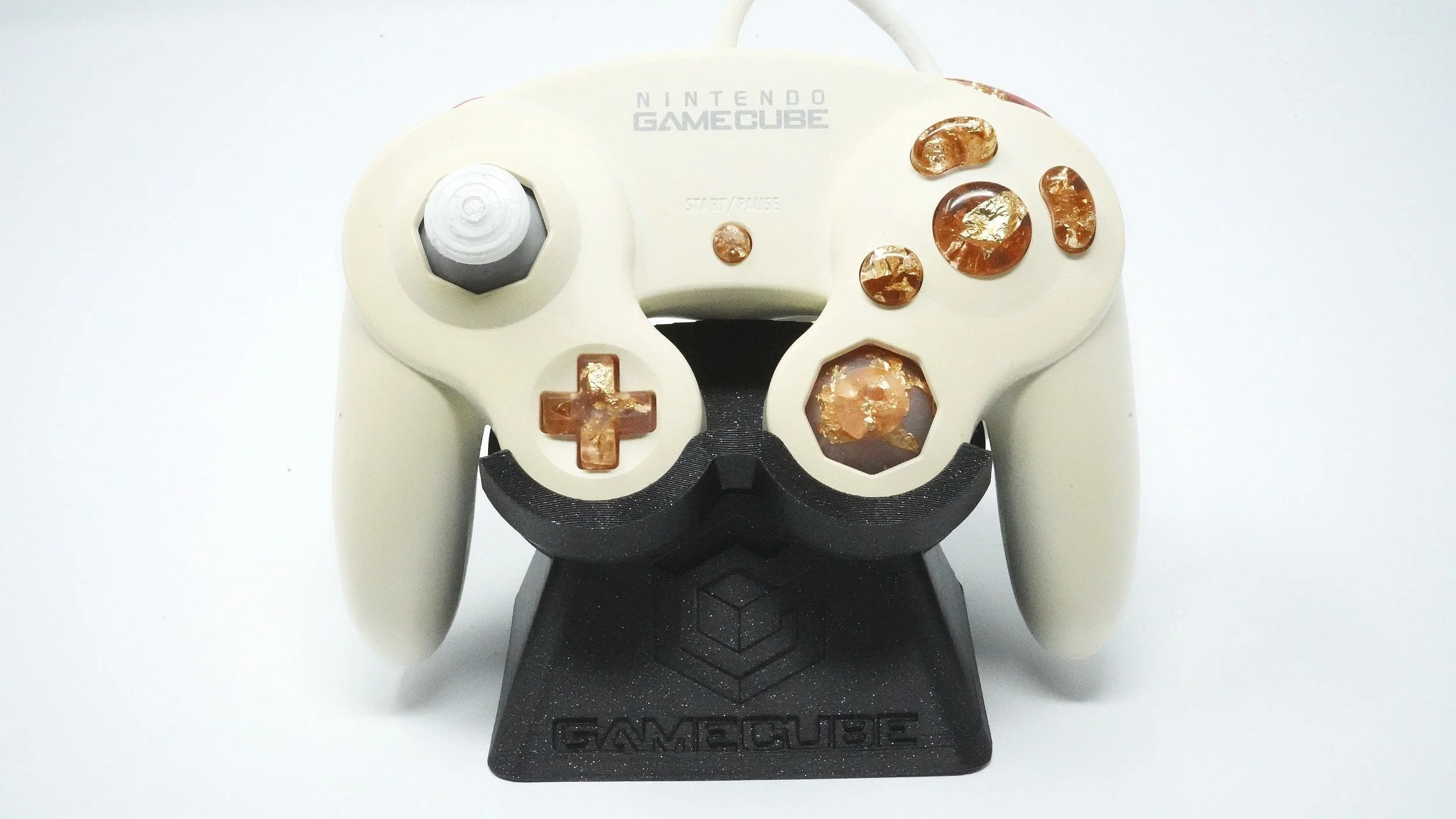 A white Nintendo GameCube controller with gold flakes inside the buttons and thumbstick, mounted on a black stand with the GameCube logo.