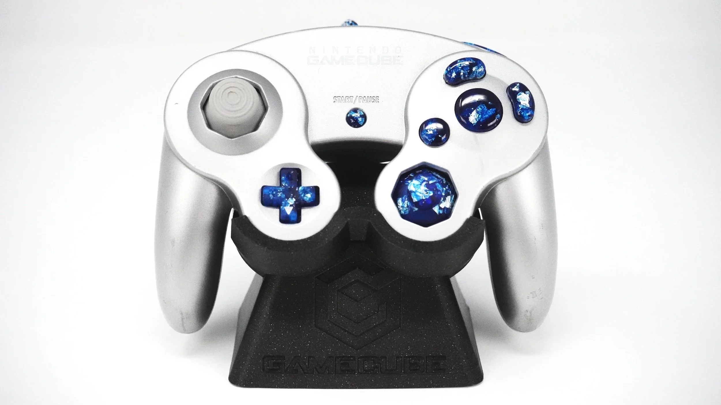 A silver Nintendo GameCube controller decorated with blue galaxy-style stickers on the buttons, with a black stand labeled 'GAMECUBE'.