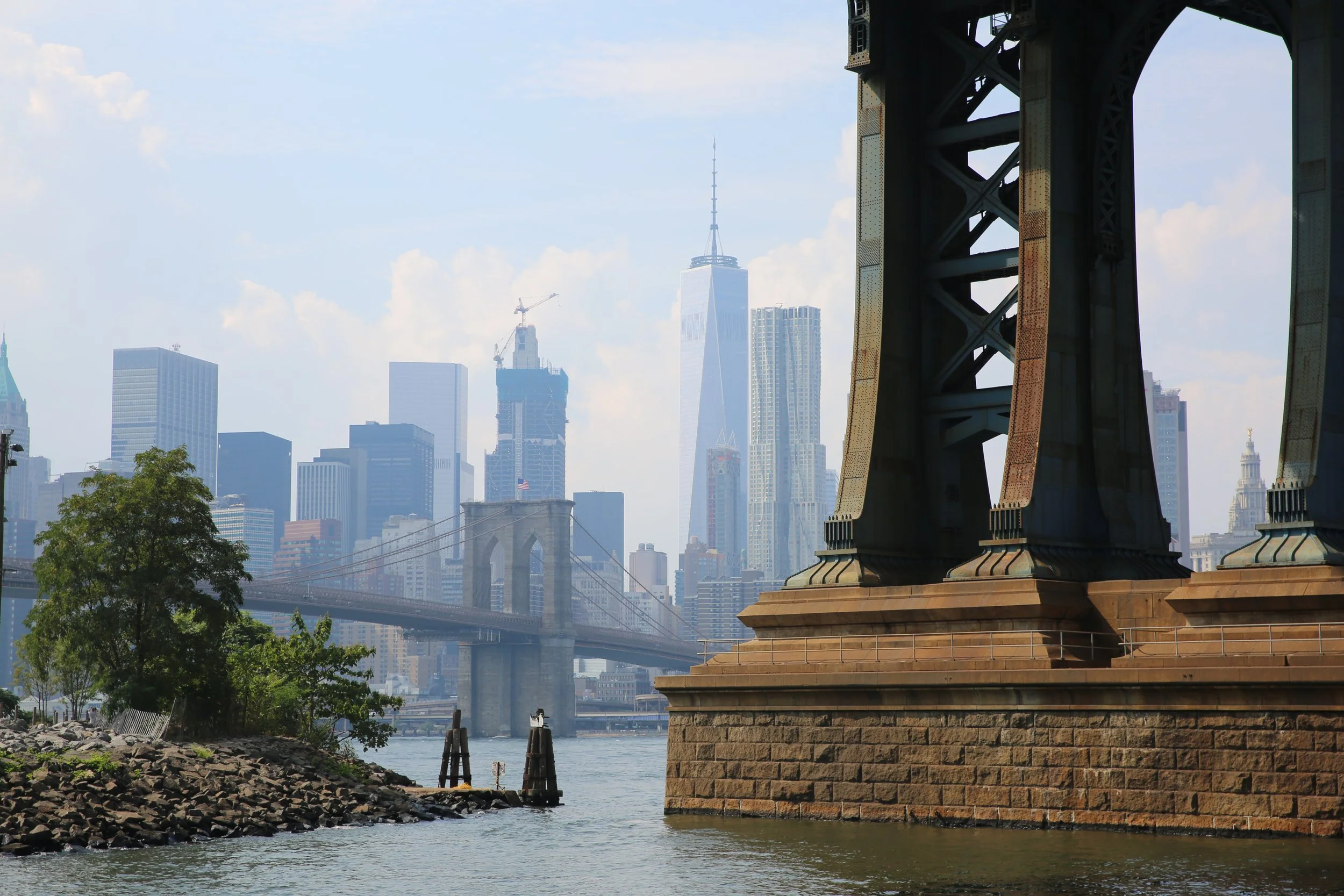 View of the New York City skyline from the Brooklyn Bridge park, showing the bridge's stone support and city skyscrapers, including One World Trade Center, under a partly cloudy sky.