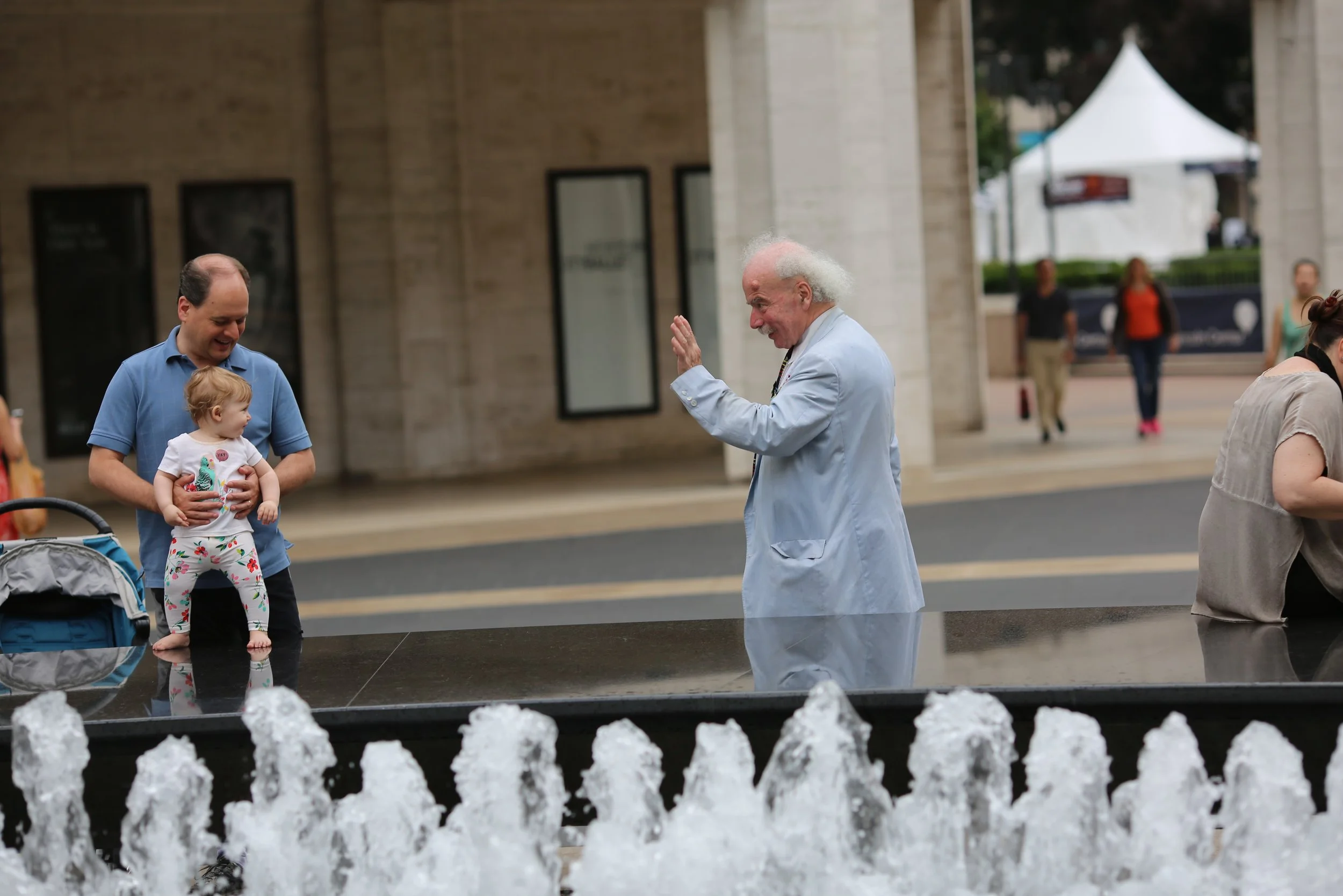A man holding a young girl near a water fountain in an outdoor urban setting. An older man in a light blue suit is smiling and waving at them. Other pedestrians are walking in the background.