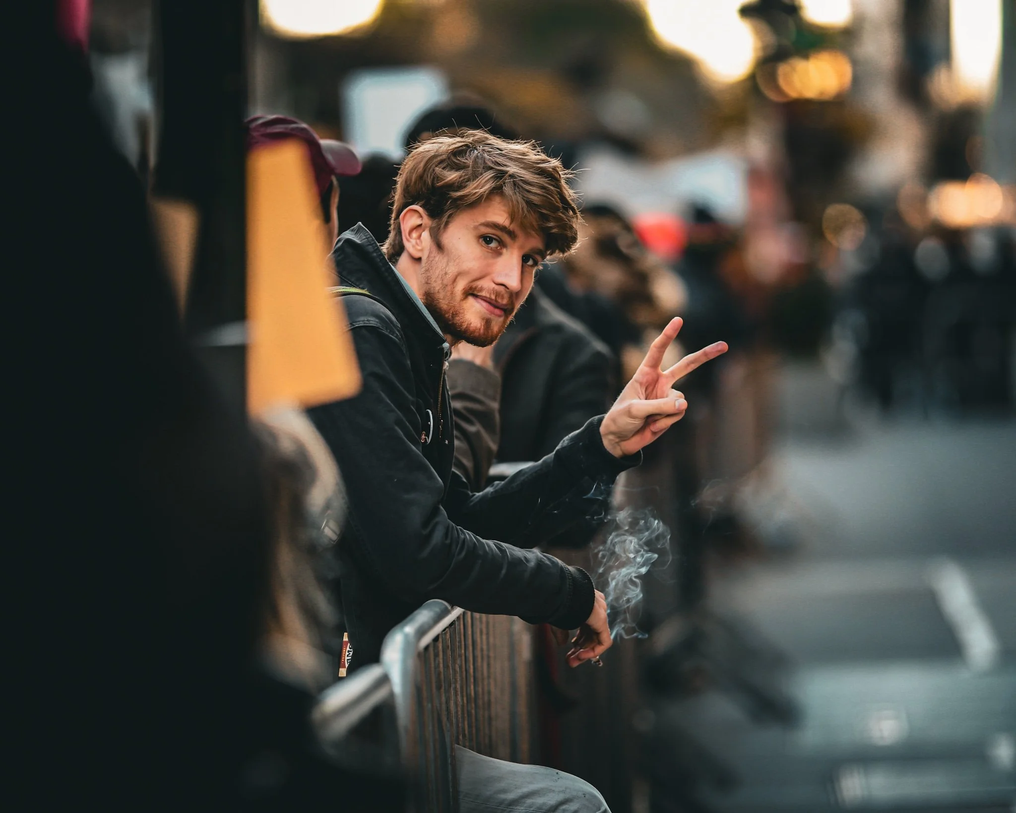 A young man with light brown hair and a beard is sitting at an outdoor location, smiling and making a peace sign with his hand. There is smoke around him, likely from a cigarette, and there are blurred people and cars in the background.