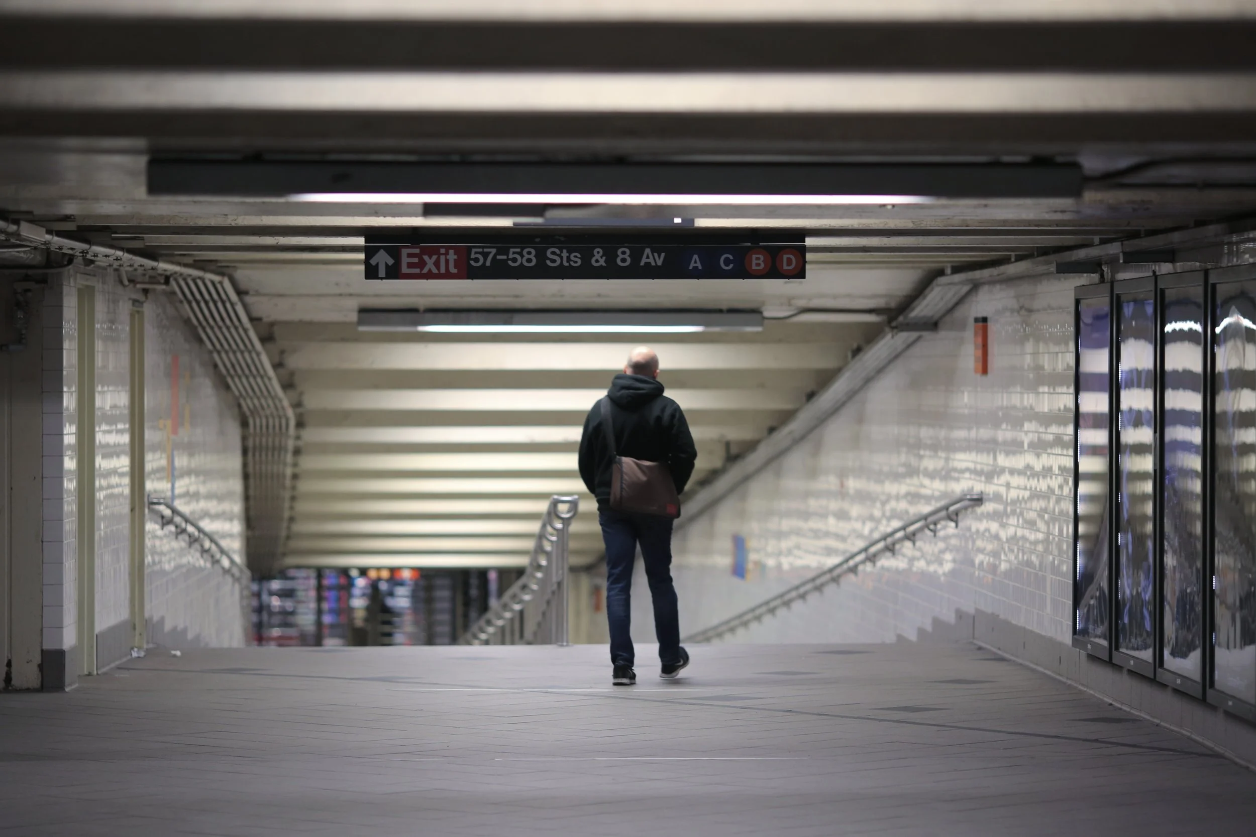 A solitary man walking in an underground subway station corridor with tiled walls and ceiling, signs indicating an exit and train lines, and glass display cases on the side.
