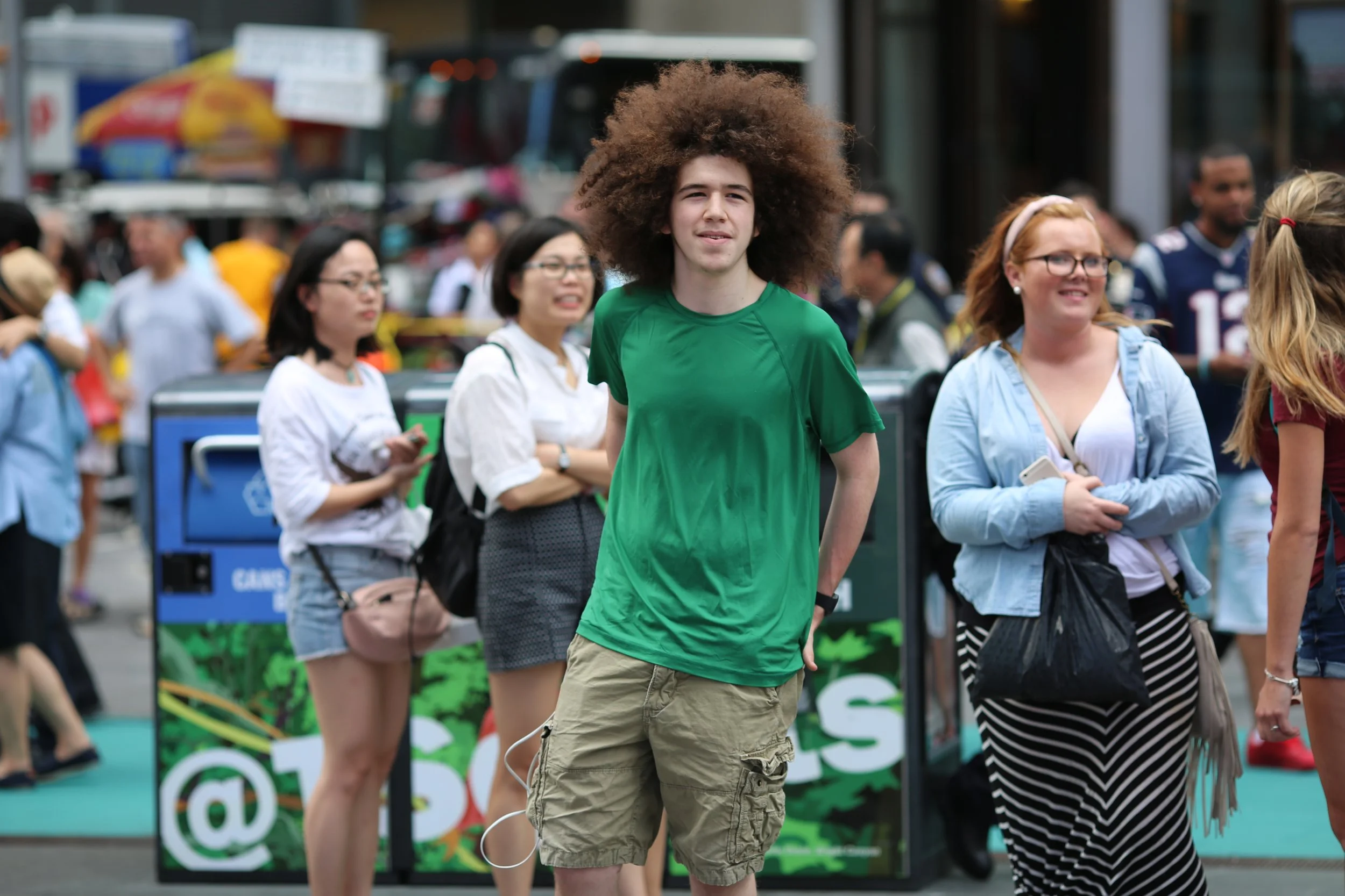 Young man with large curly hair wearing a green T-shirt and khaki cargo shorts standing on a busy city street surrounded by pedestrians.