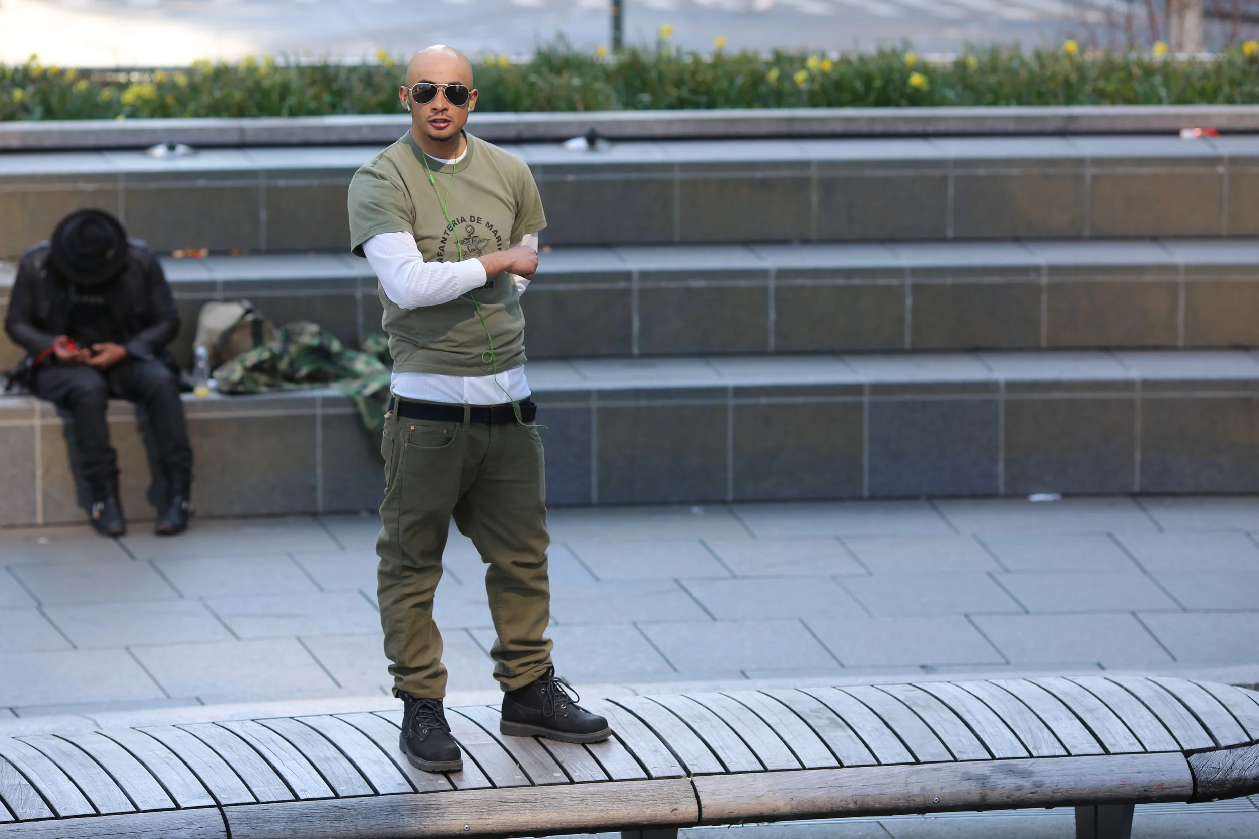 A man wearing sunglasses, a green t-shirt, and olive cargo pants standing on a curved wooden bench in an urban setting, with a man in black sitting in the background.