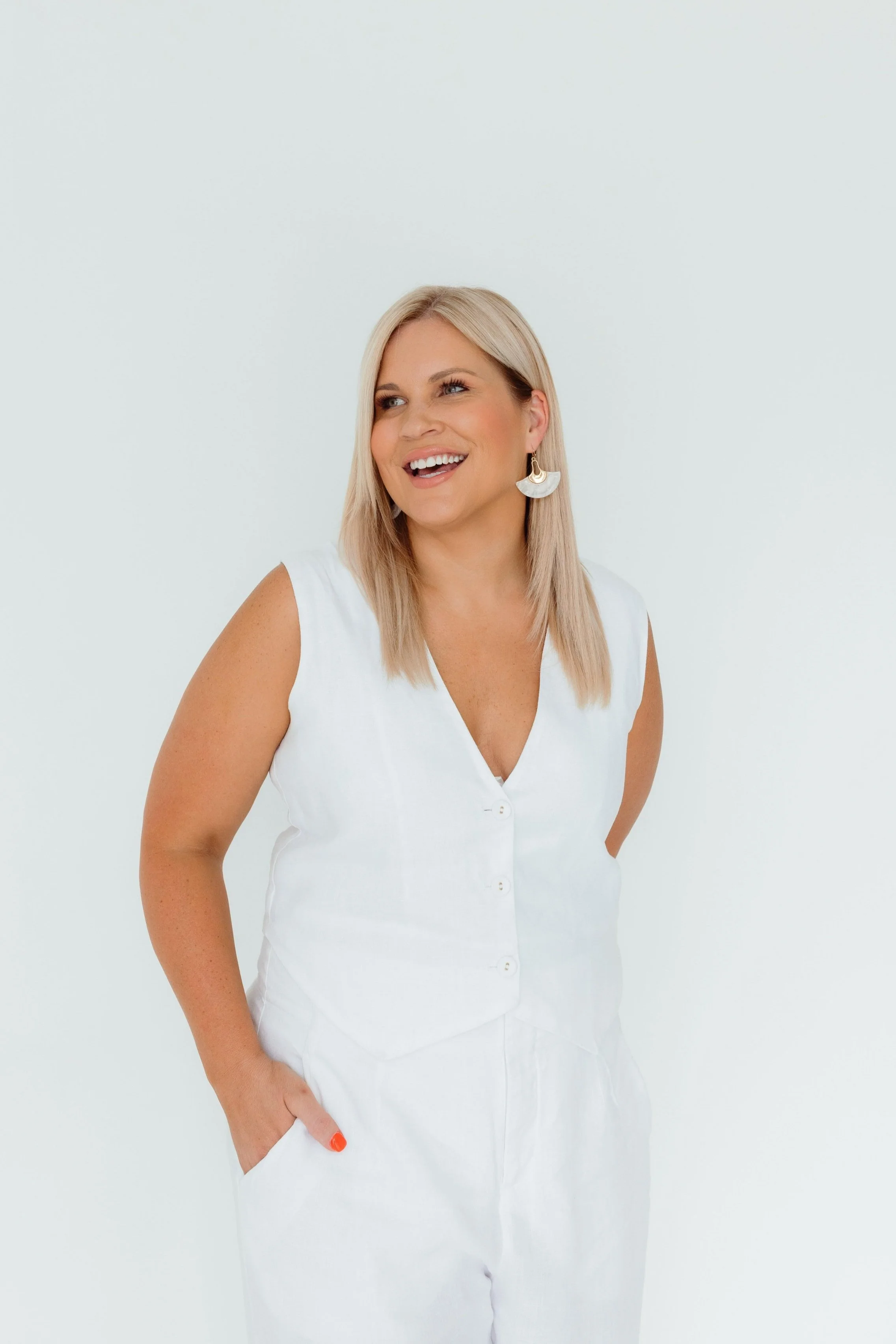 A woman with blonde hair wearing a white sleeveless button-down top and matching white pants, smiling and standing against a plain white background.