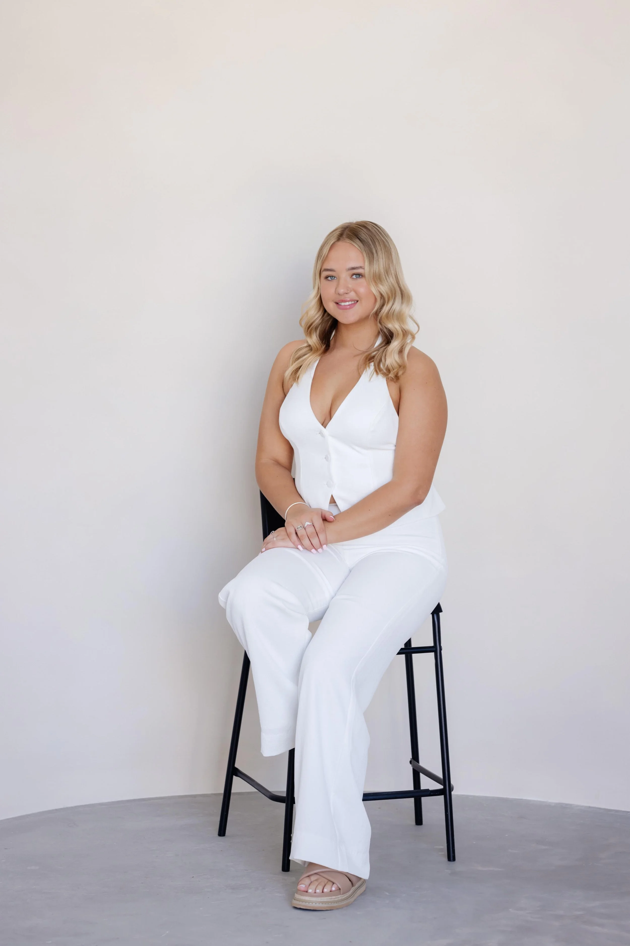 A young woman with blonde wavy hair, wearing a white sleeveless top and white pants, is sitting on a black stool against a plain white background, smiling at the camera.