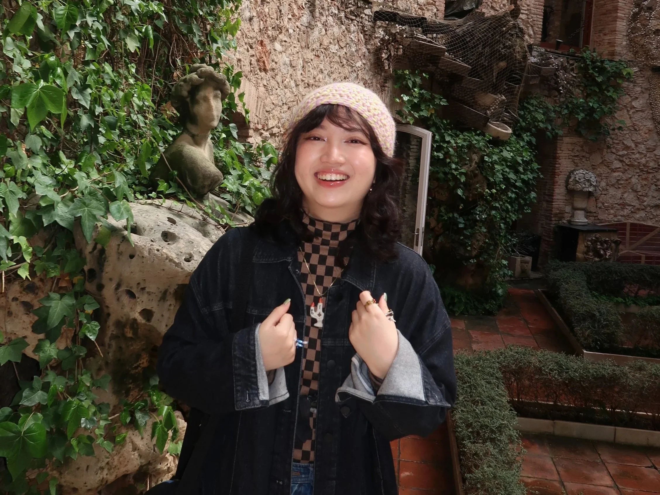 A young woman with dark hair wearing a pink knit beanie, black denim jacket, checkered top, and rings, smiling and looking at the camera inside a courtyard with stone walls, green ivy, a bust sculpture, and potted plants.