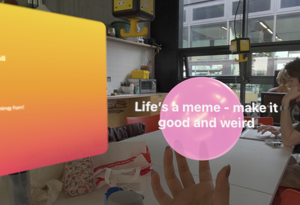 A person sitting at a table in a modern office or coworking space, using a laptop, with a pink translucent bubble over their hand displaying the words 'Life's a meme - make it good and weird.' There are other people working, large windows, and hanging yellow schedule boards in the background.