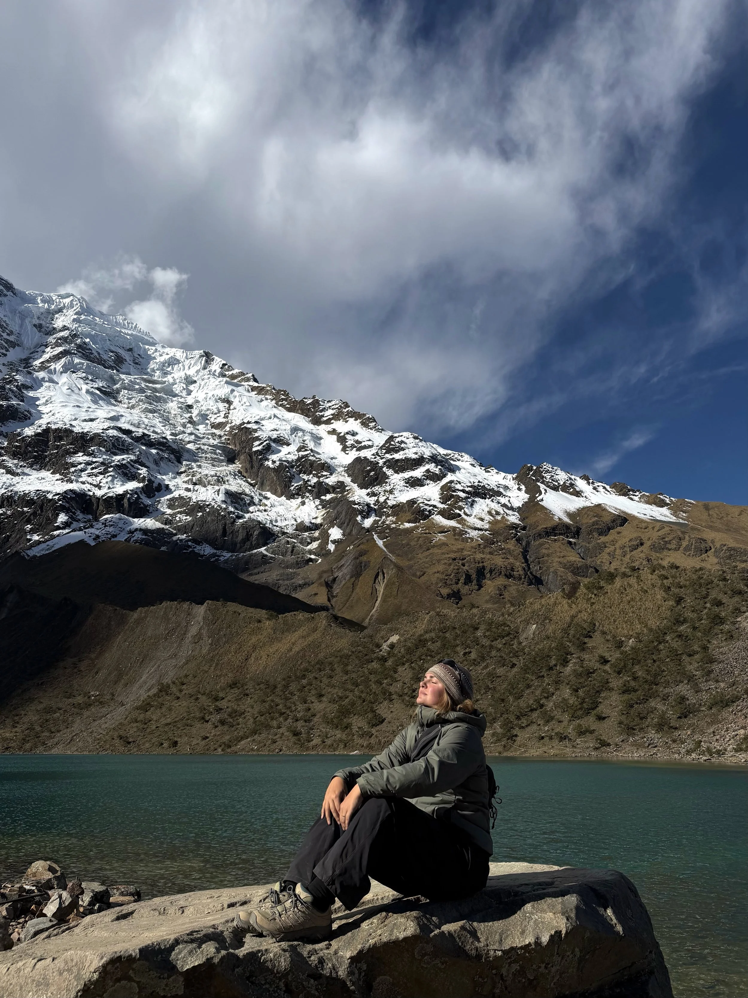 Maddie sitting by a turquoise alpine lake, eyes closed in the sun