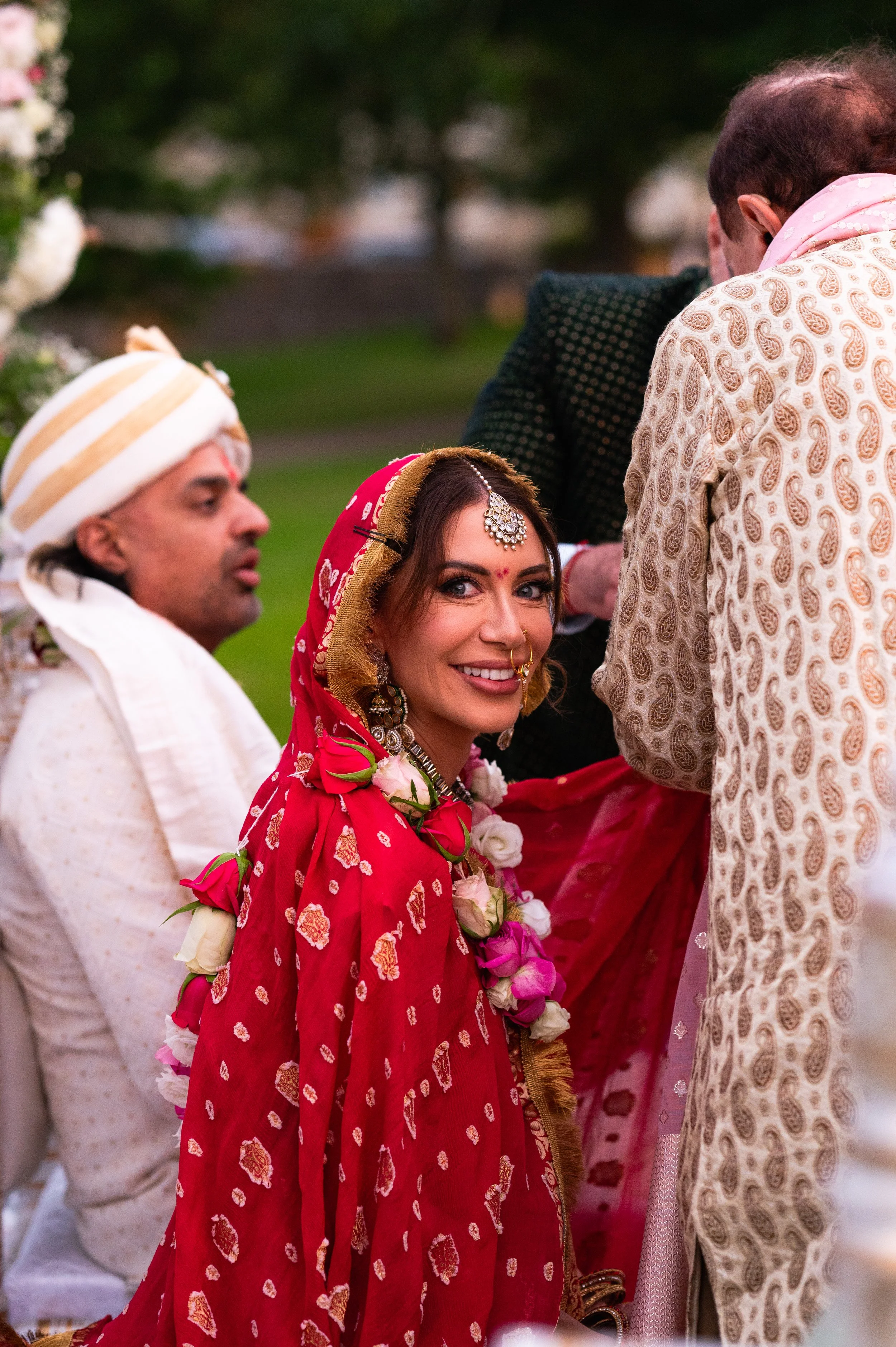 A woman dressed in traditional Indian wedding attire, smiling and surrounded by family during a ceremony.