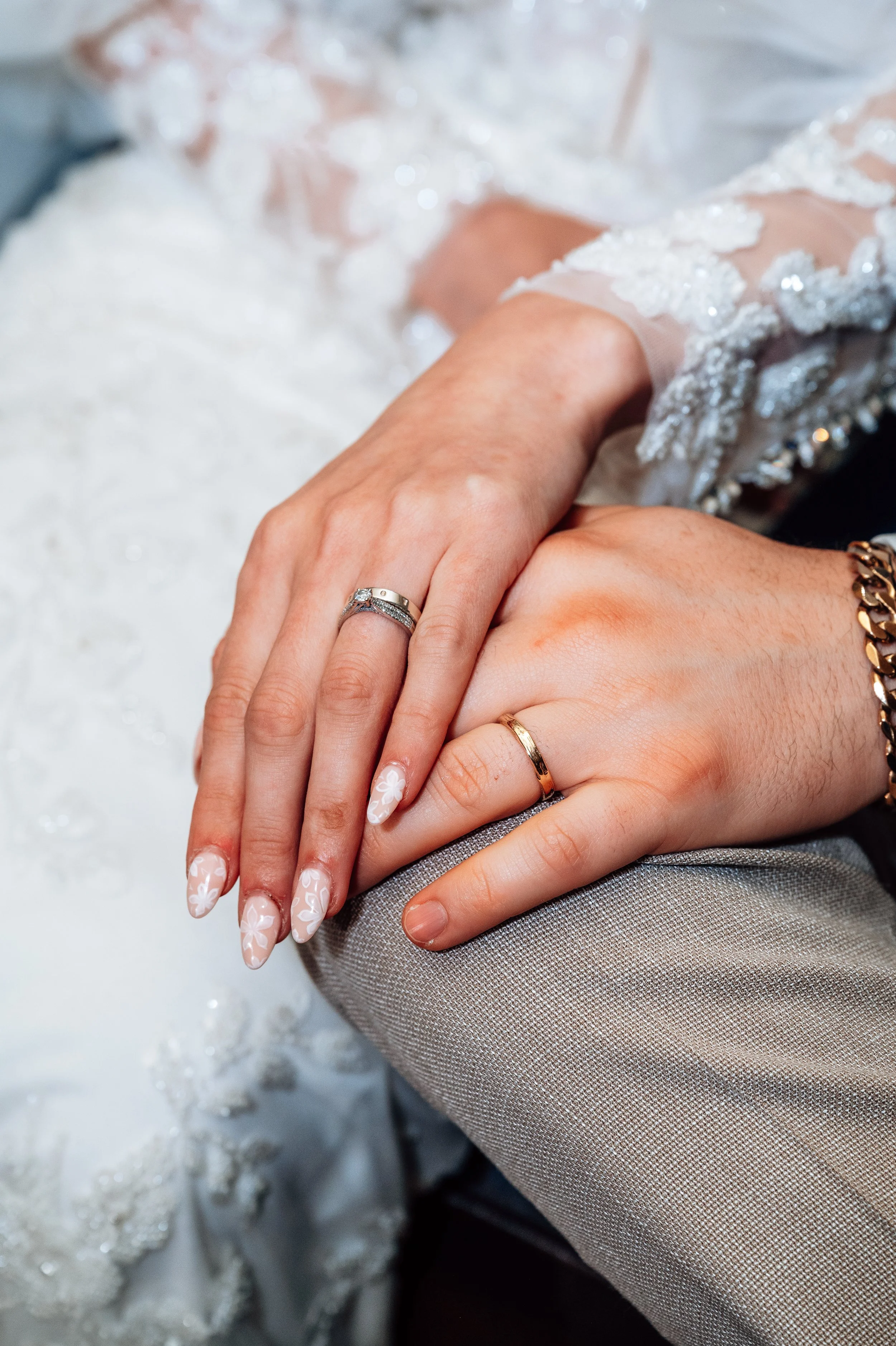 Close-up of a bride and groom's hands with wedding rings, resting on the bride's lap. The bride's nails are decorated with white floral nail art and she wears a delicate ring and a gemstone ring. The groom's hand has a simple gold wedding band and a chain bracelet. The bride is in a lace wedding dress and the groom is wearing a textured suit.
