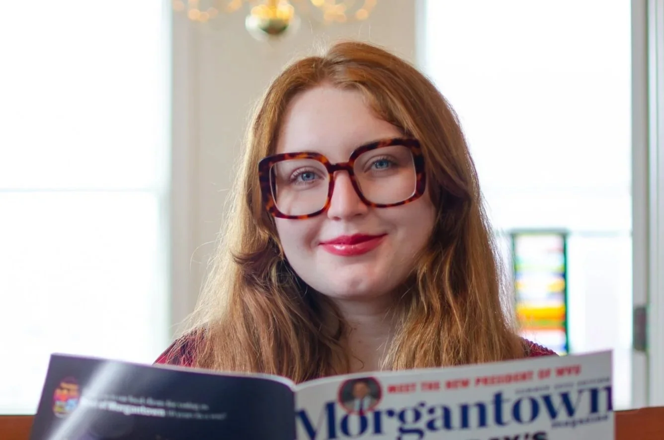A red-haired woman with large tortoise-shell glasses smiling while reading a newspaper.