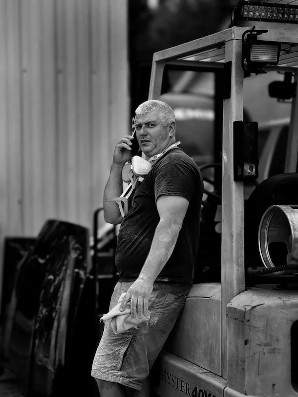 A man is leaning against a construction vehicle while talking on a cellphone. He is holding a cloth in his left hand and has a supply mask around his neck.