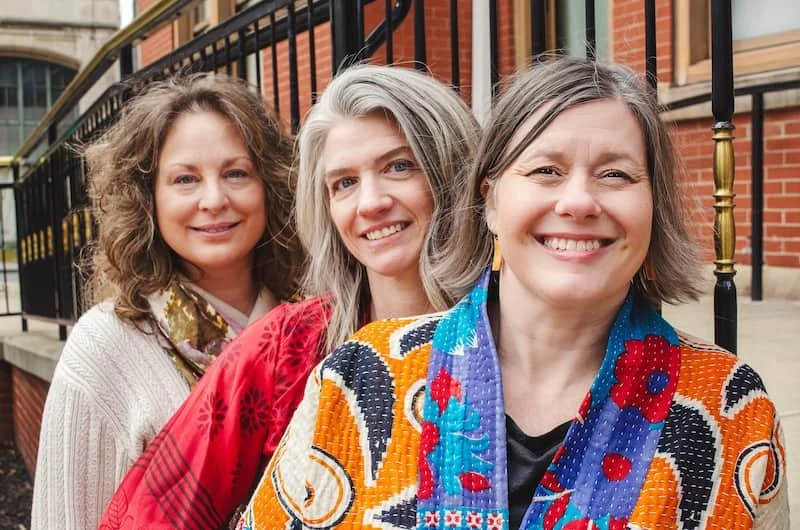 Three women standing outdoors in front of a building with a black fence, smiling at the camera.