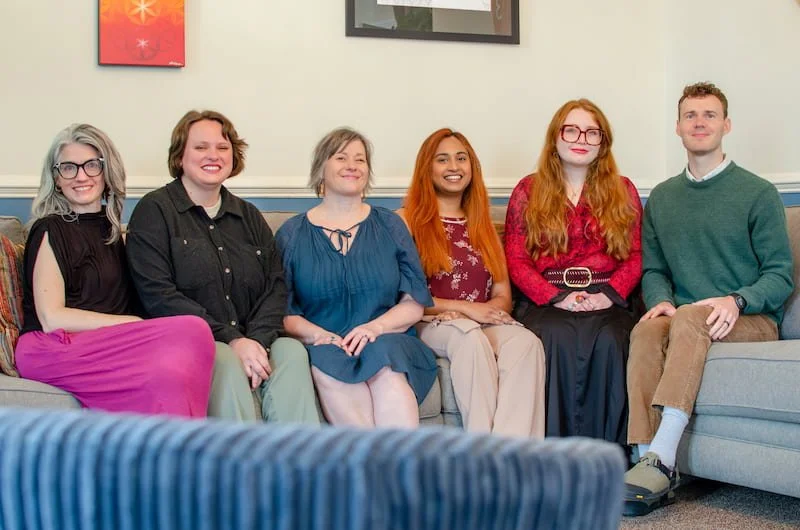 Six people sitting on a beige couch in a living room, smiling for a group photo. The background features white walls with two framed artworks.