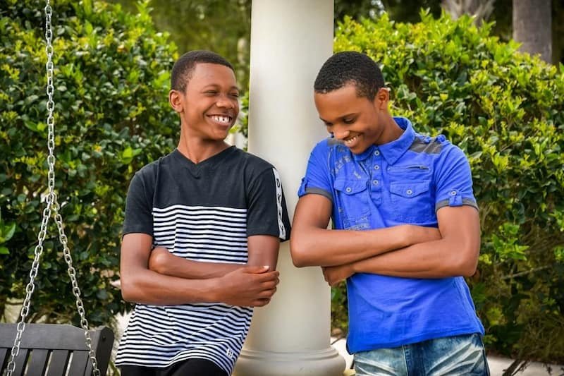Two boys standing outdoors near a white column, smiling and talking with arms crossed.