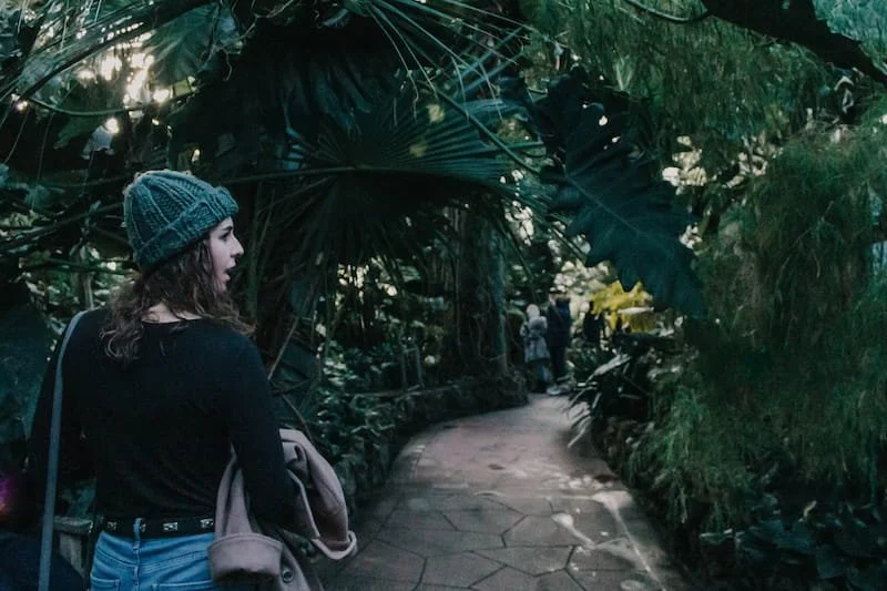 A woman wearing a gray knit hat and black shirt carrying a tan bag on a winding path in a lush, tropical garden, with other visitors in the background.