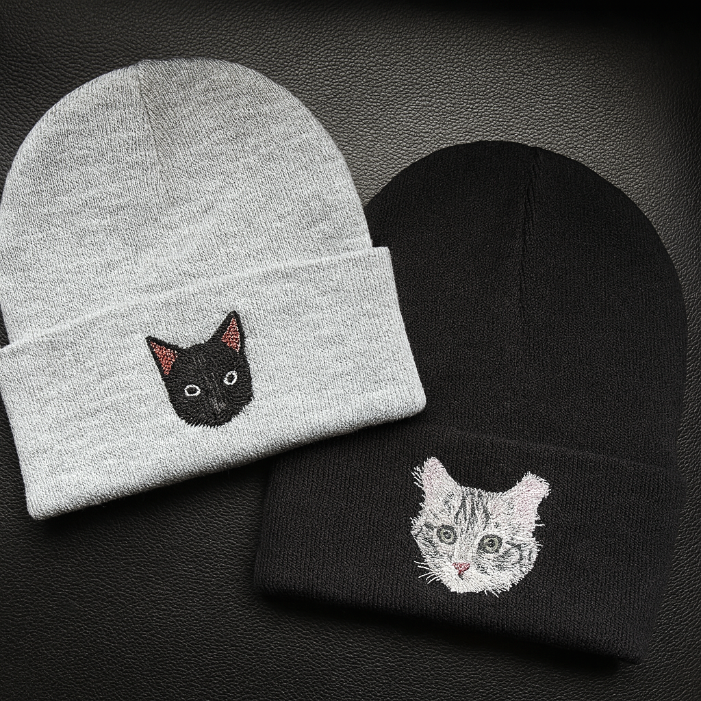 Two knit beanies, one gray with a black cat face embroidery, and one black with a gray tabby cat face embroidery, on a black surface.