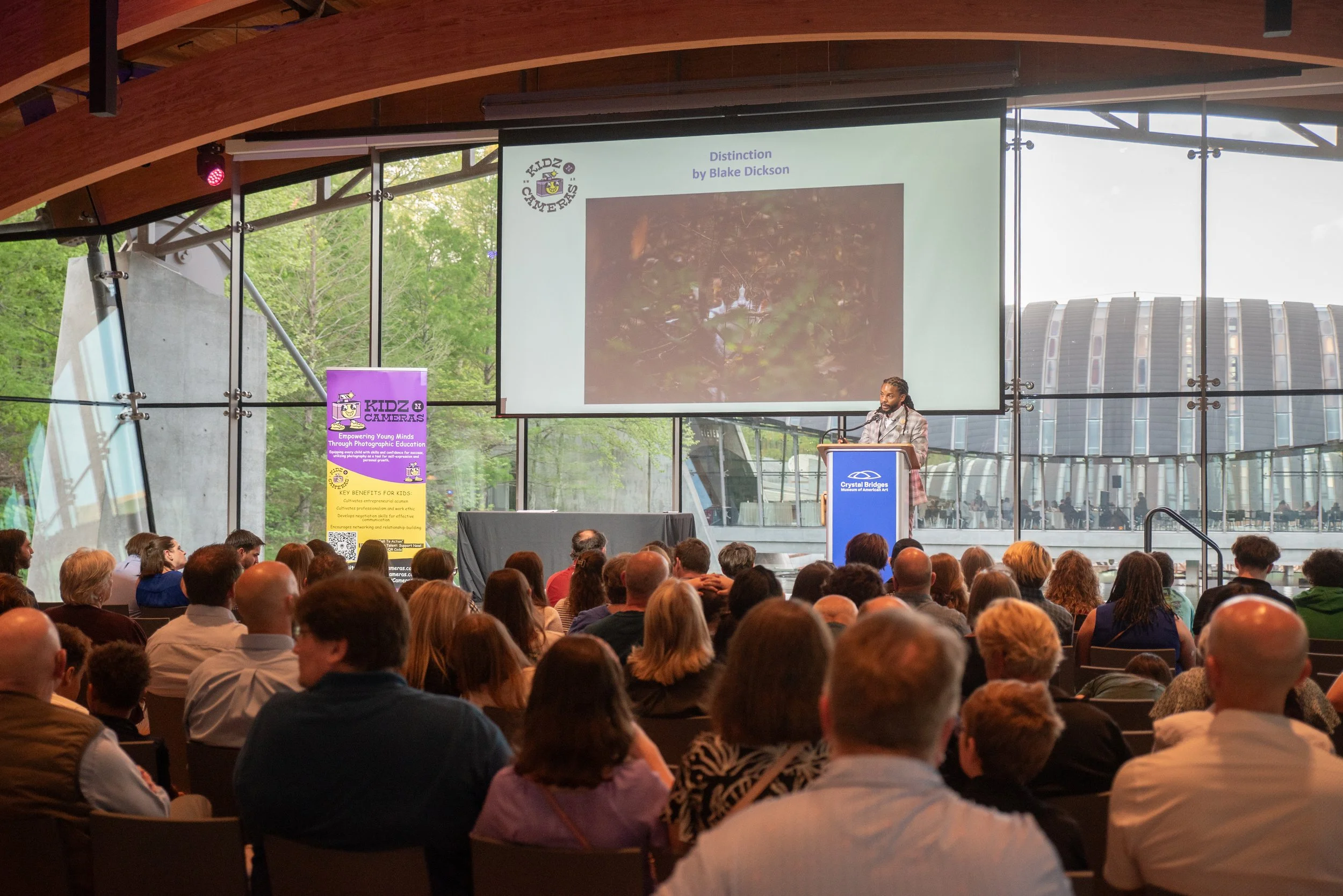 A speaker on stage at a conference or event with a large audience. The stage has a screen displaying a presentation slide titled "Distinction" by Blake Dickson. There are banners promoting Kidz Cameras and the Crystal Bridges Museum of American Art.