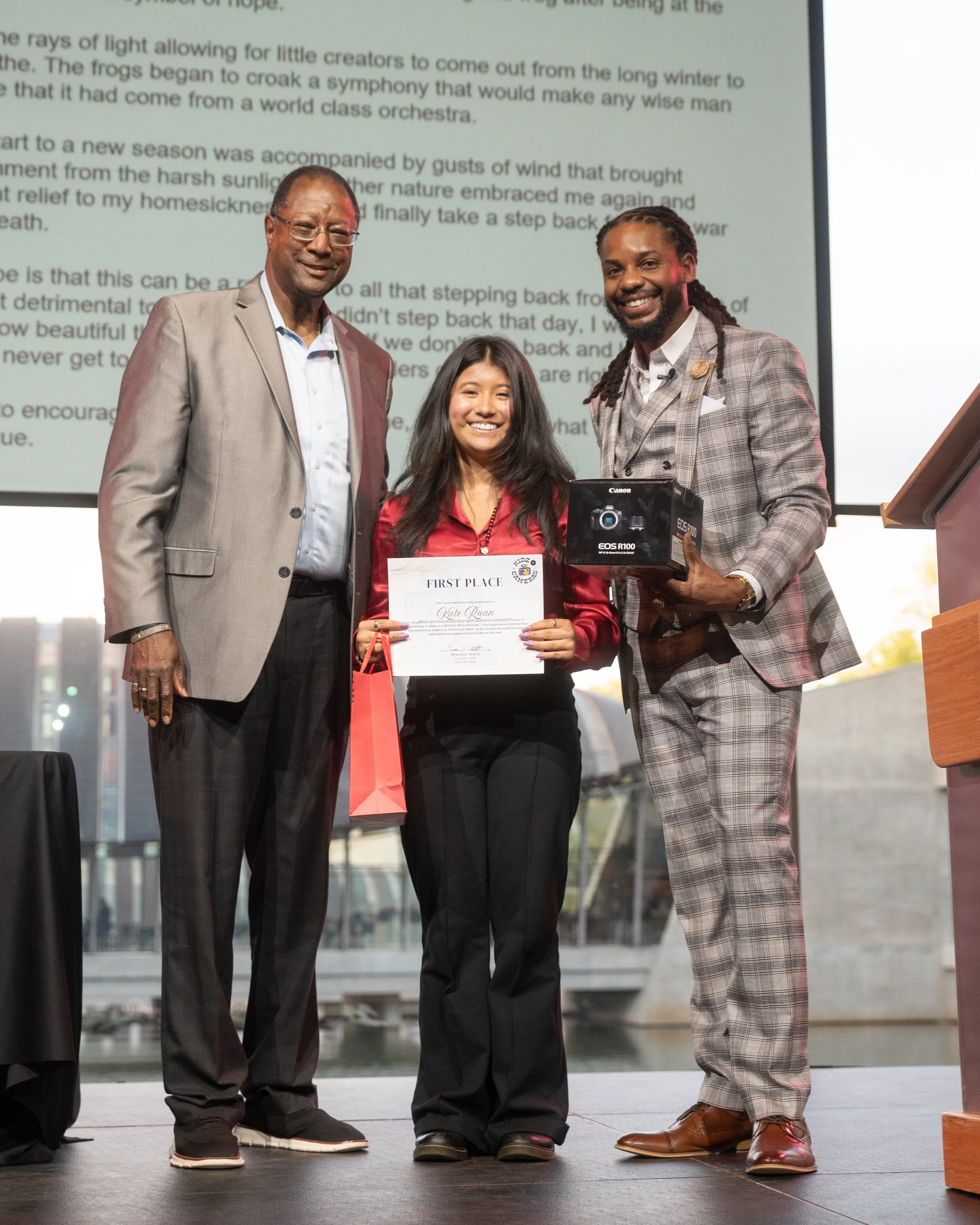 Three people standing on stage during an award ceremony, with a large screen displaying text behind them. The person in the middle is holding a certificate that says 'First Place,' and a red gift bag. The person on the right is holding a Canon EOS R100 camera box. The scene appears to be indoors with natural light coming through large windows.