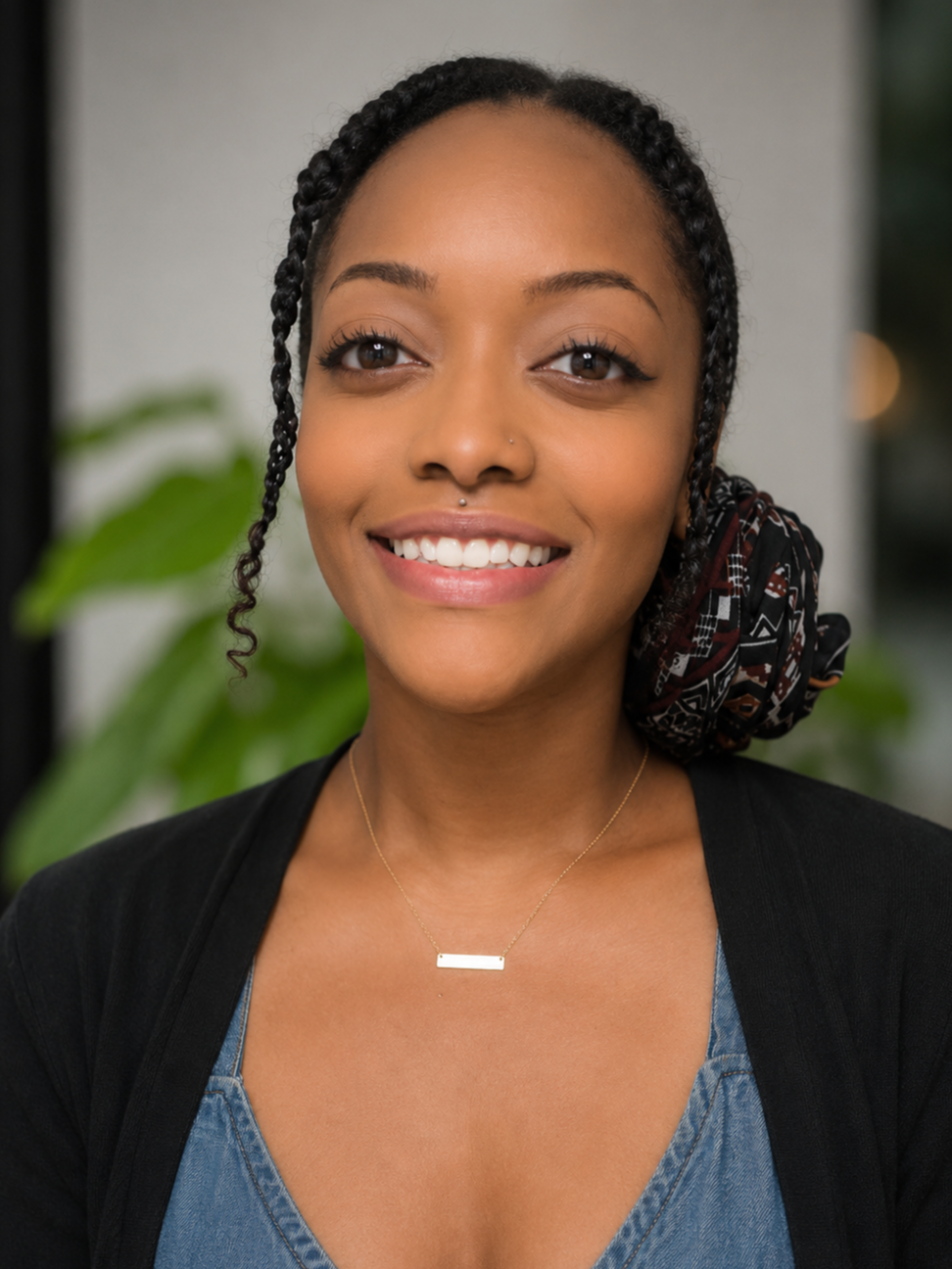 A young woman with braided hair, smiling, wearing a black top, denim shirt, a necklace, and a patterned scarf tied around her hair in a bun, indoors with green plants in the background.