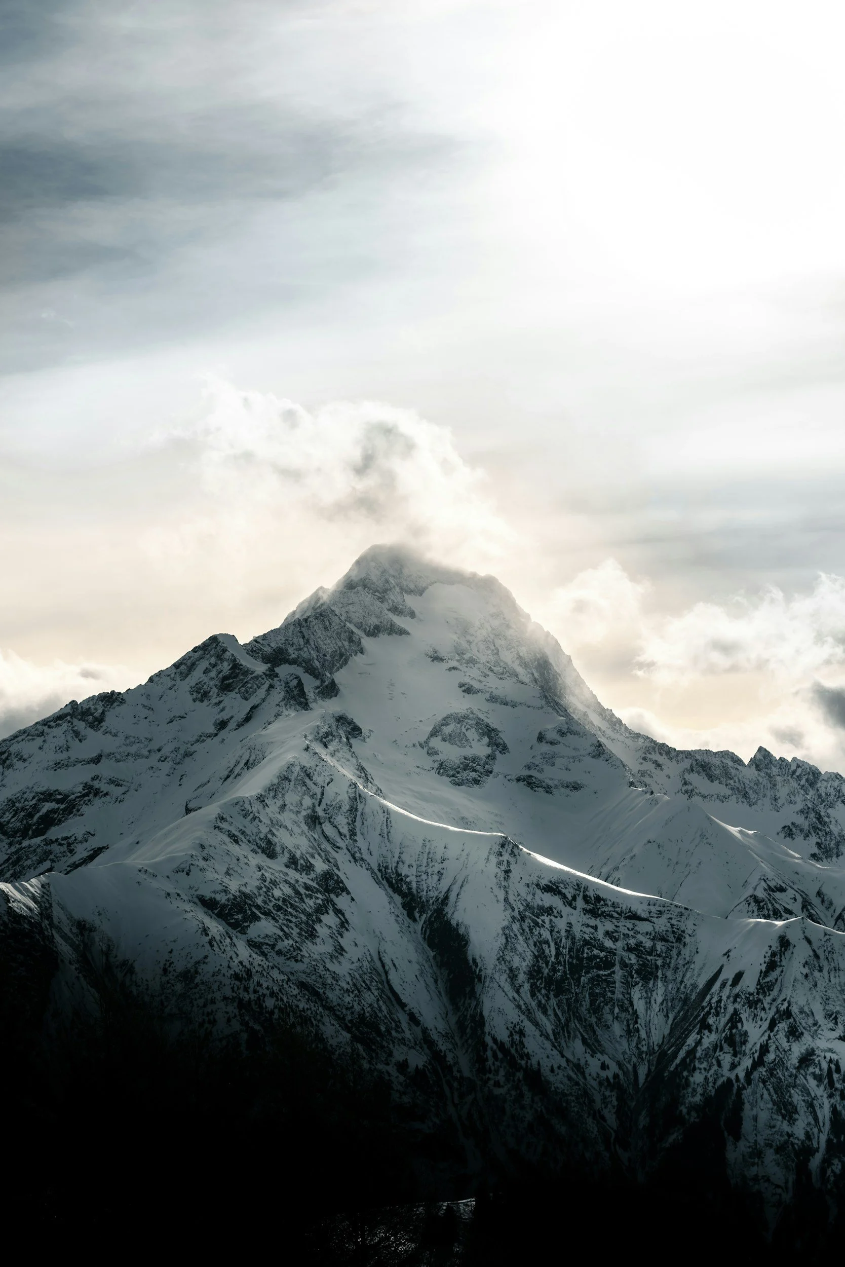 Close up of a snow covered mountain peak with light cloud cover and bright shining light..