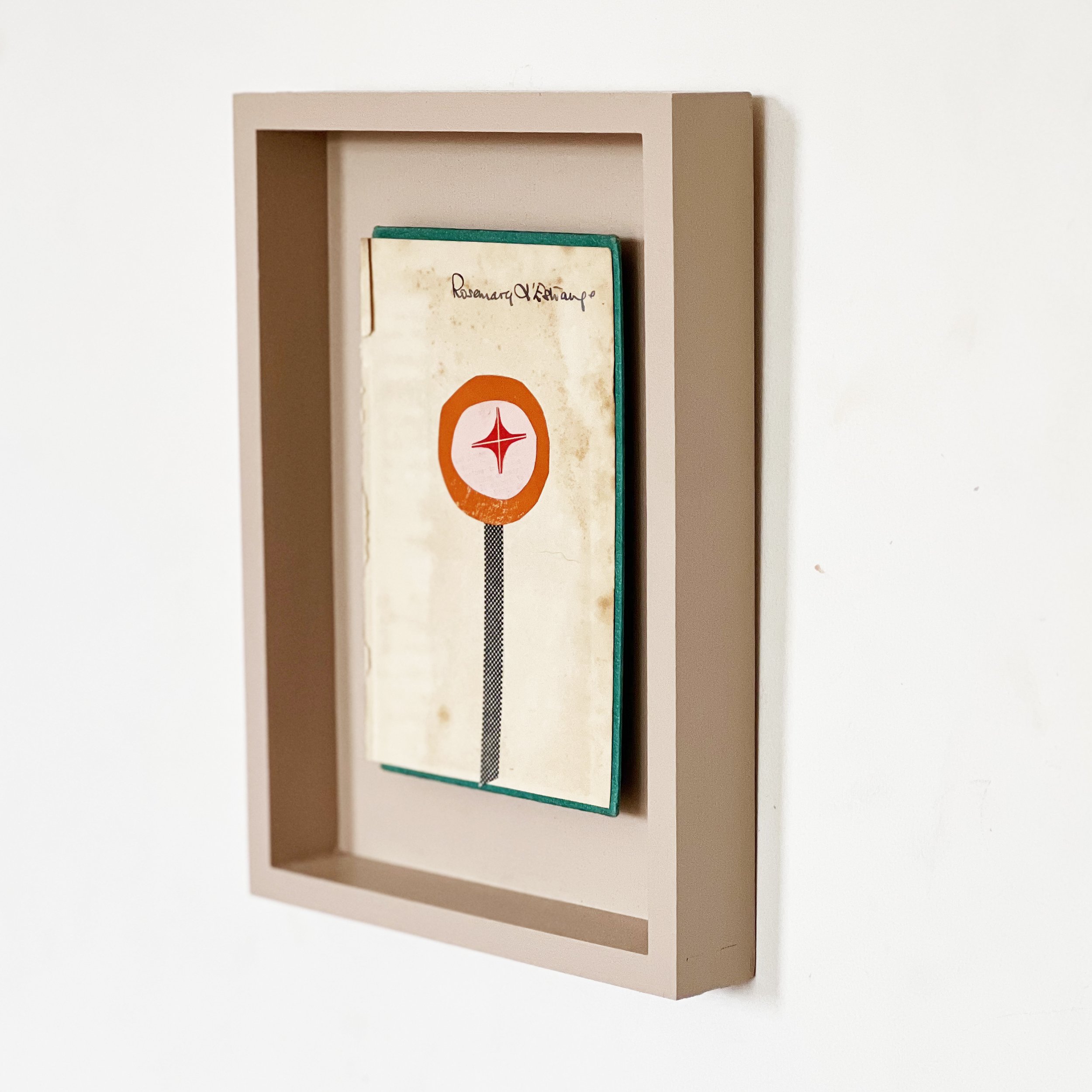 A collage artwork depicting a small orange flower with a red star in the centre. Created on a worn vintage book cover shown from a side angle.