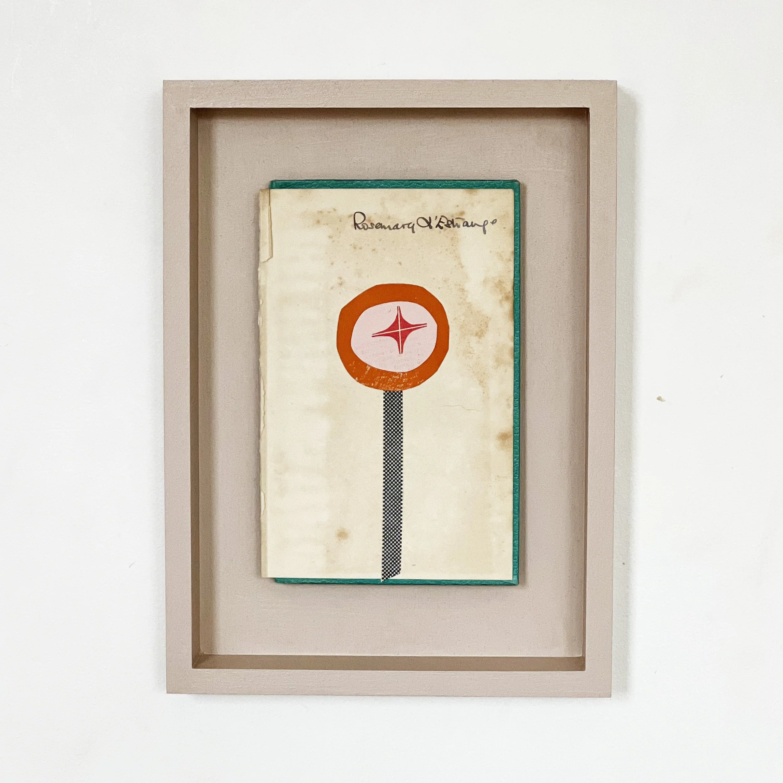 A collage artwork depicting a small orange flower with a red star in the centre. Created on a worn vintage book cover and mounted within a pale pink open frame.