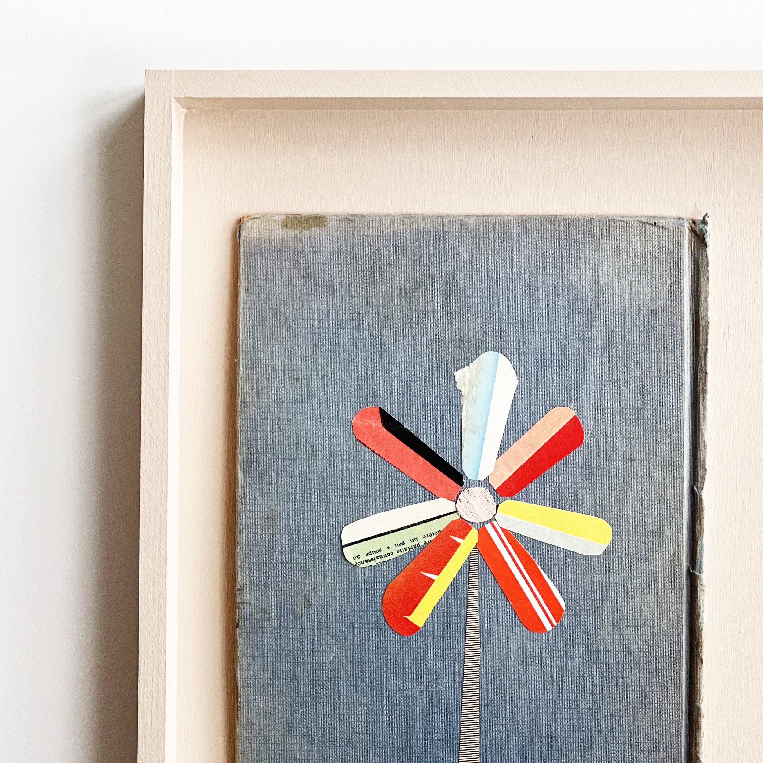 A collage artwork depicting a flower with colourful petals. Created on a worn blue vintage book cover and mounted in a pale pink open frame.