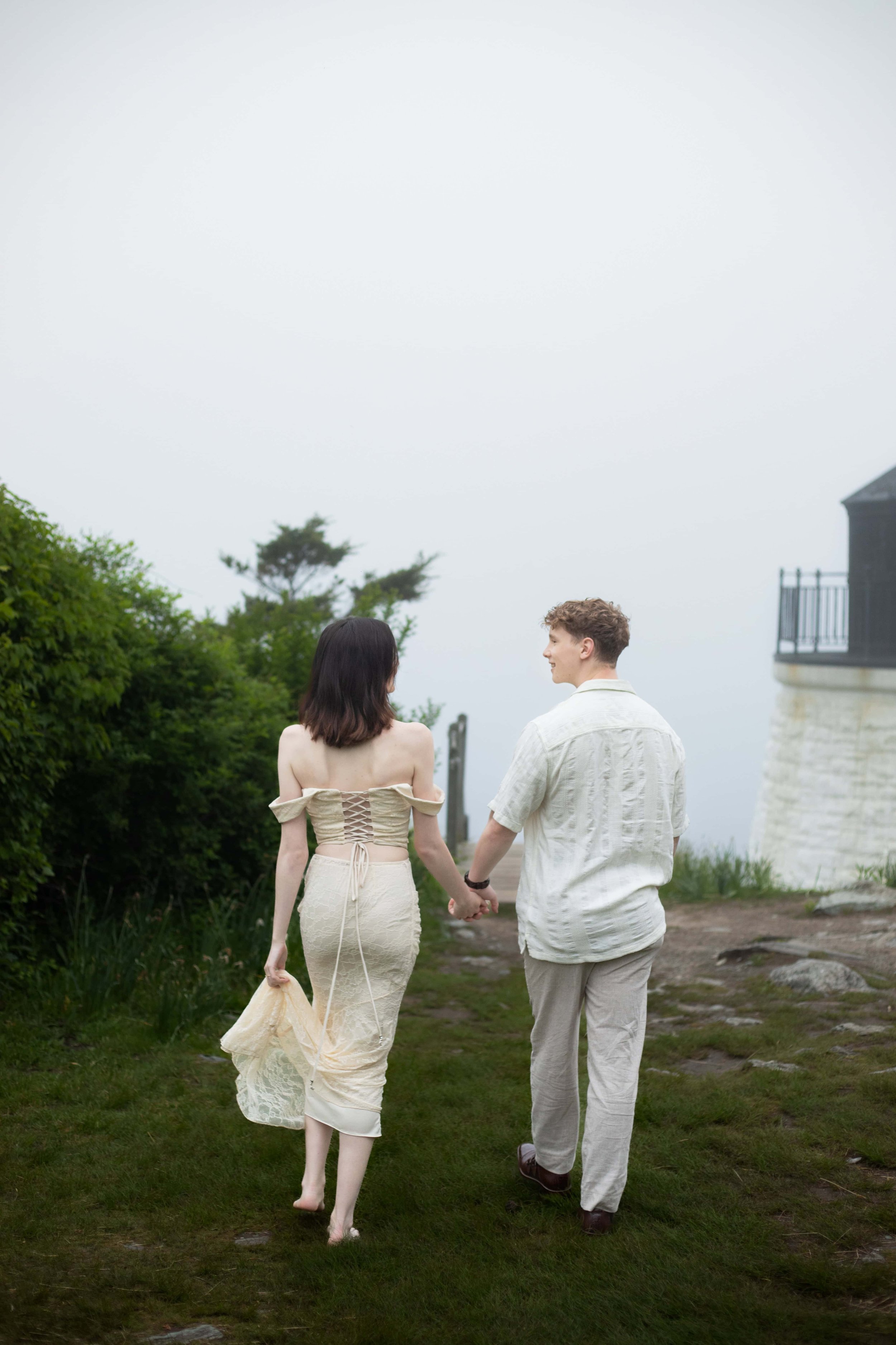 Newly engaged couple is walking hand in hand toward the stairs near the lighthouse at Castle Hill Inn. She is holding the bottom of her dance in her hand.