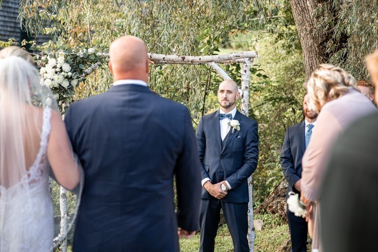 Groom holding tears back at bride walks down the aisle at an outdoor ceremony at Jones River Trading Post