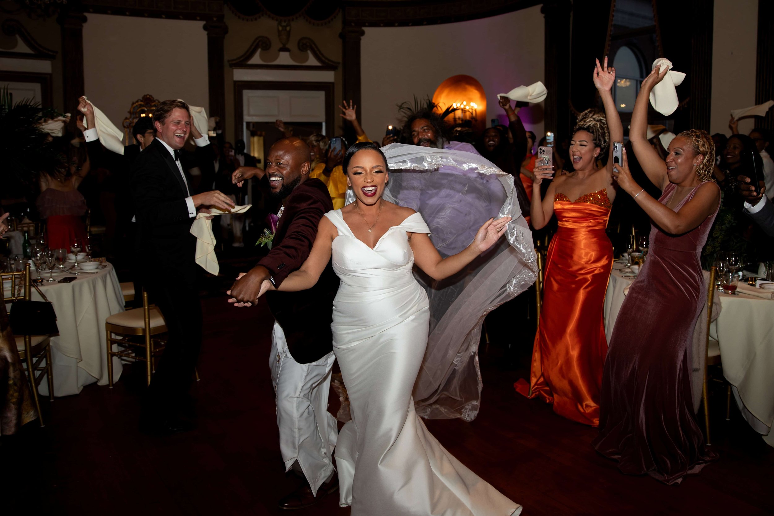Bride and groom walking into their reception at the Graduate in Providence, RI. Bride is wearing a simple white dress and has her veil on flying in the wind behind her as she runs in. Groom is wearing traditional Ugandan traditional wedding attire.