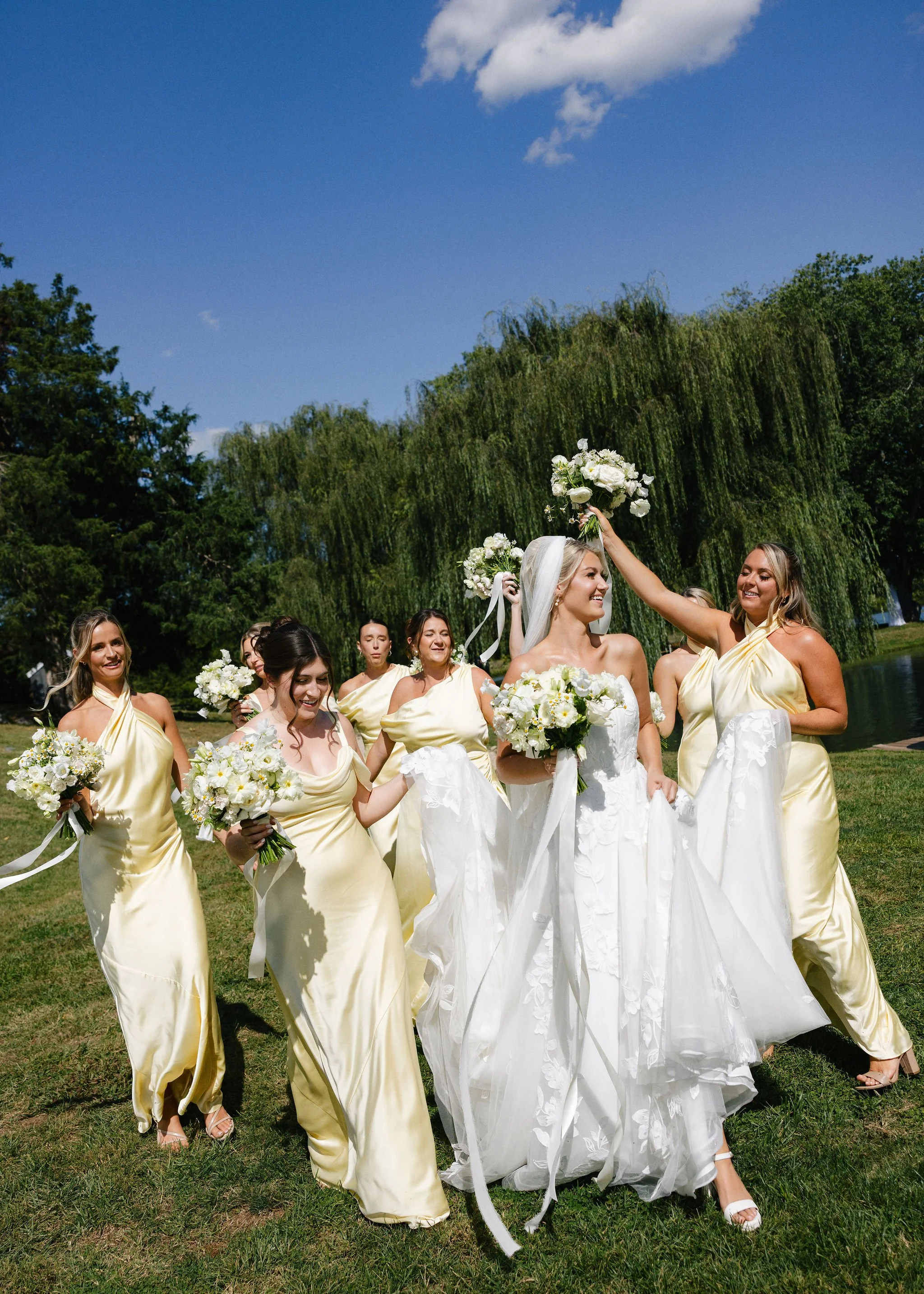 Bride and bridesmaids walking in butter yellow bridesmaids dresses on lawn with blue sky