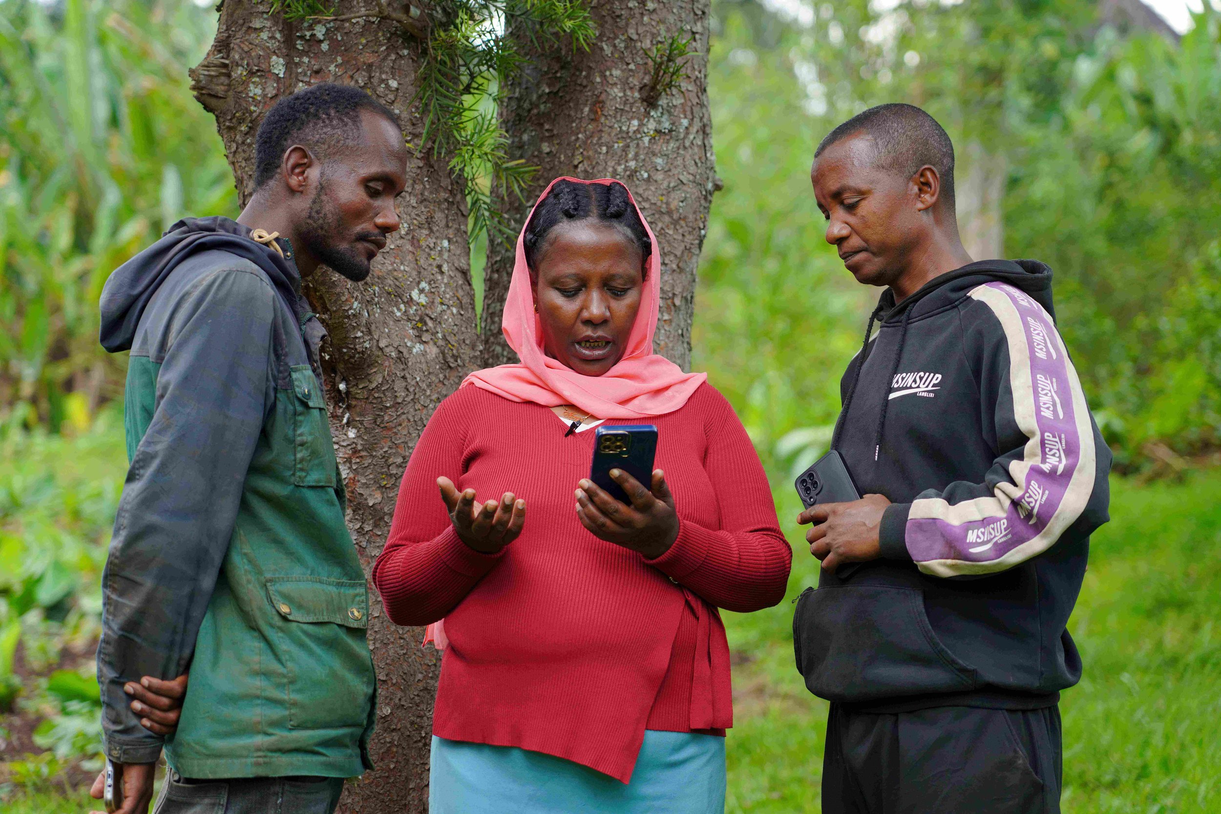 A woman in a red sweater and headscarf holds a smartphone while a man stands beside her outdoors during a Digital Green Farmer.Chat program visit.