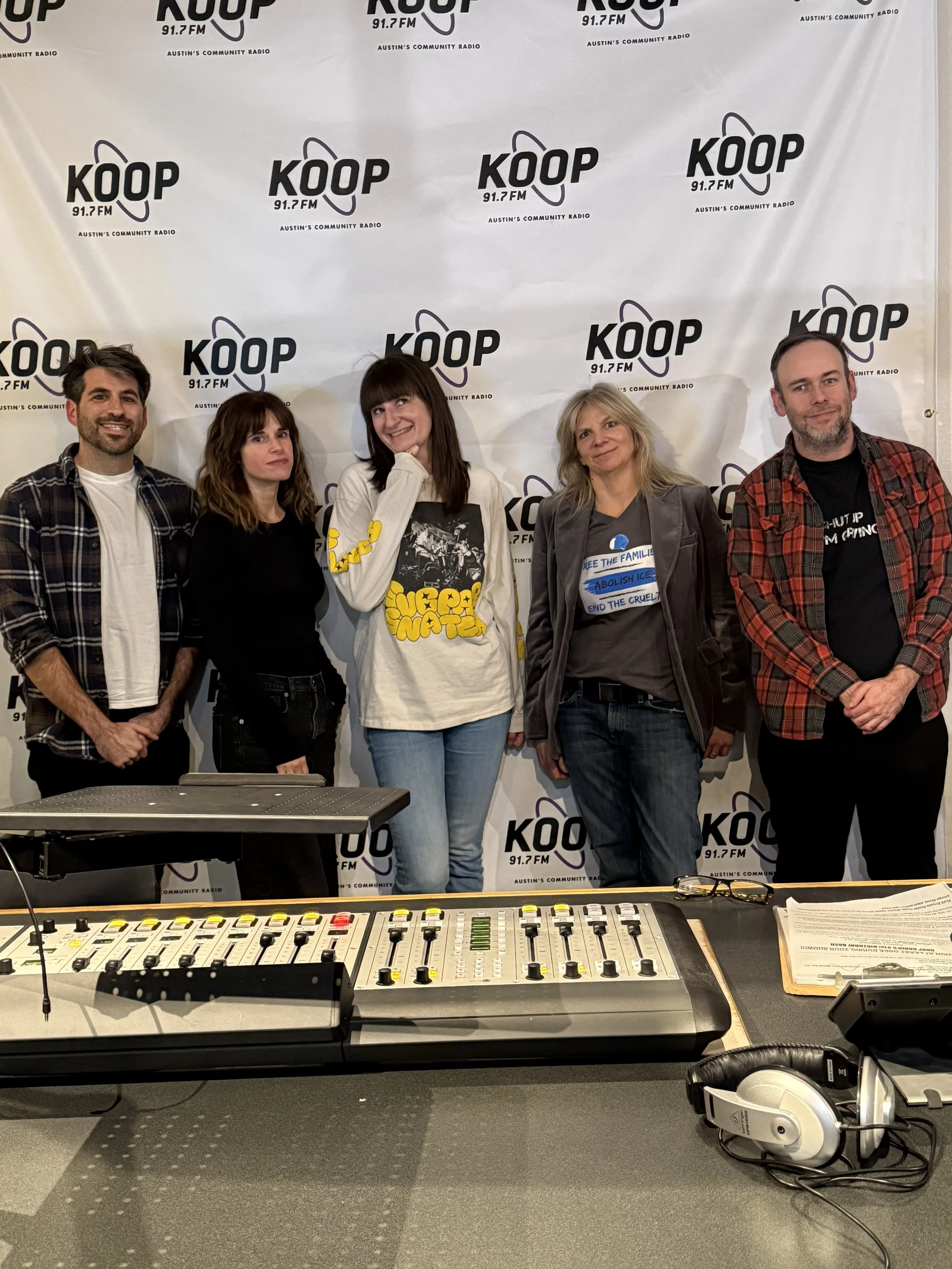 Five people standing in front of the KOOP 91.7 FM DJ Booth in Austin, Texas. On the left is DJ Cameron. On the right is the Austin-based band, Wild Heaven. From left to right, Wild Heaven is Aja Pollock, Laura Blewitt, Andi LaGrone, and Blake Bowles.