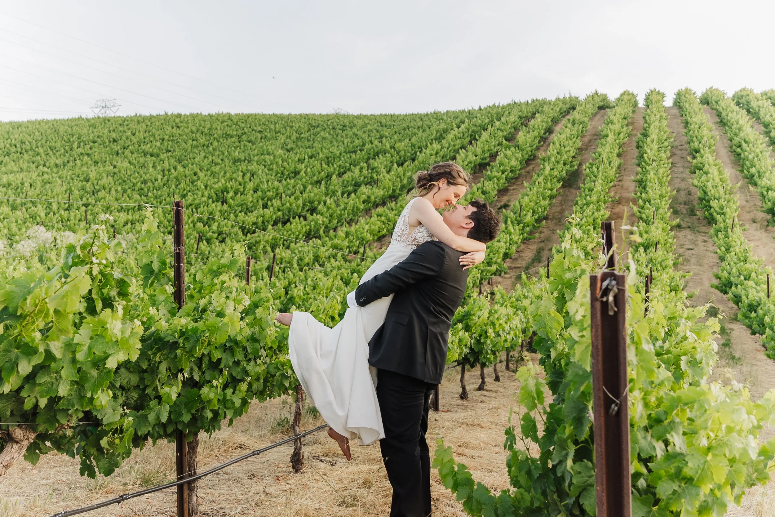 A couple, with the woman in a white dress and the man in a black suit, are embracing in a vineyard, with the woman being carried by the man, both smiling and looking at each other.