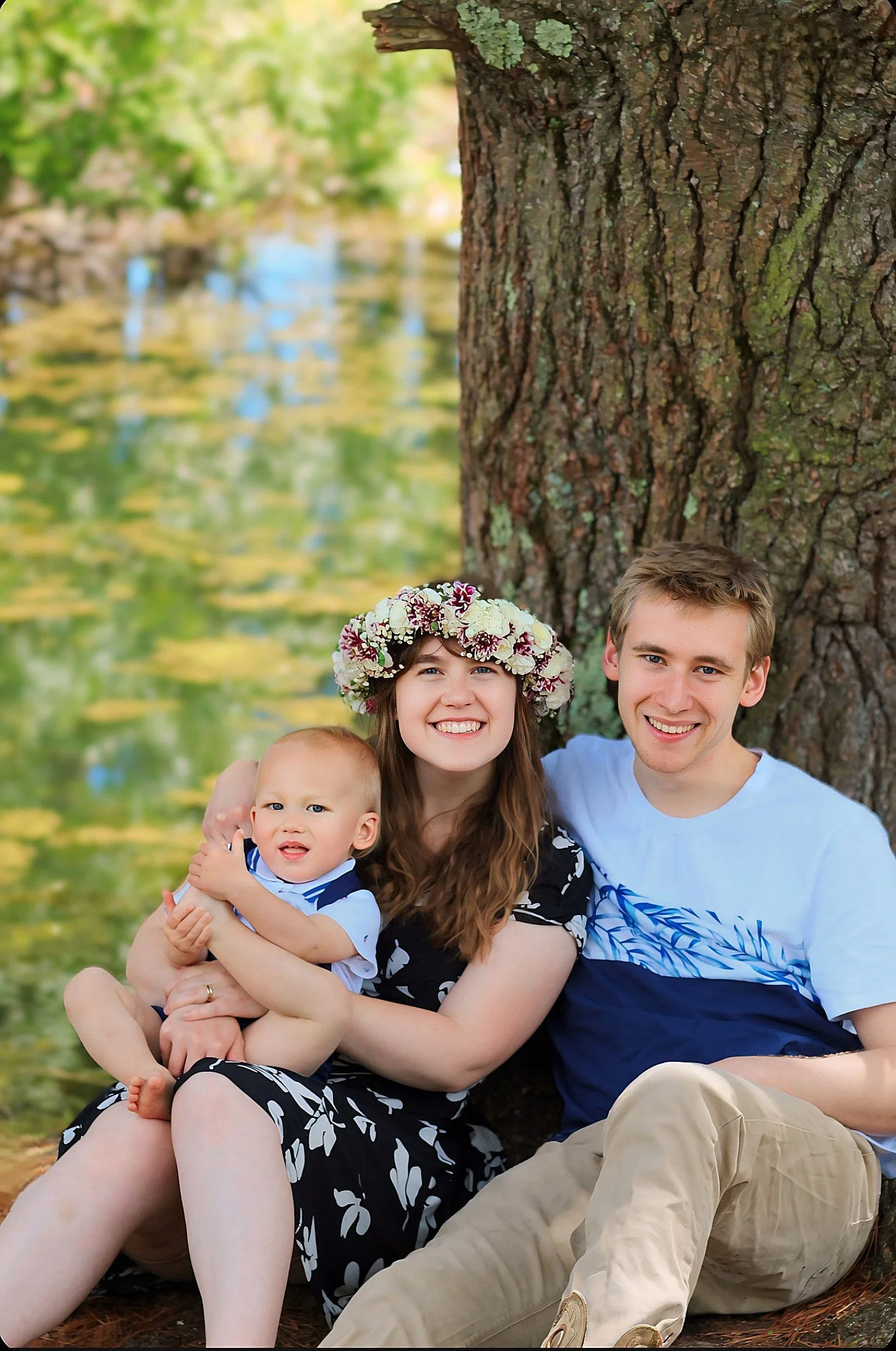A family of three sitting outdoors near a tree with a lake in the background. The woman wears a flower crown, the man is in a white and blue shirt, and a baby is in her lap.
