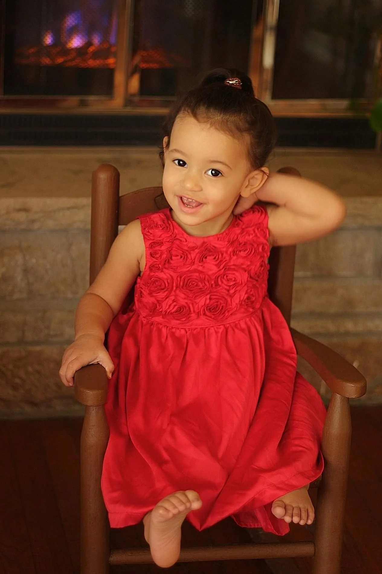 A young girl sitting on a wooden chair, wearing a red dress with a floral pattern on the top, smiling, with her right hand resting on her head and her left hand on the armrest. In the background, there appears to be a fireplace with a fire burning.