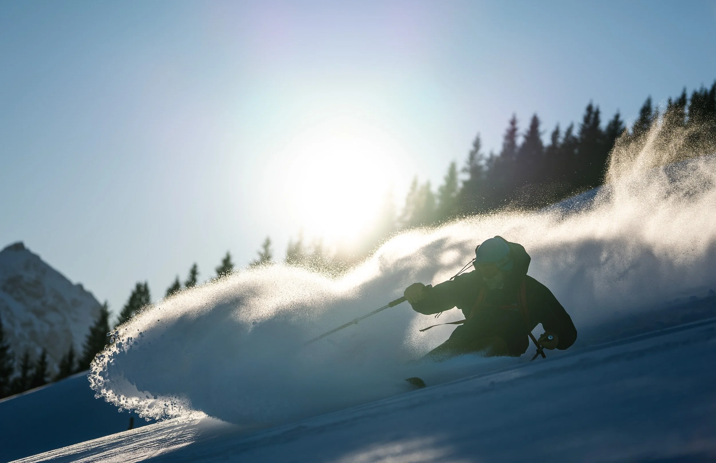 A person skiing on a snow-covered mountain with pine trees and a bright sun in the background