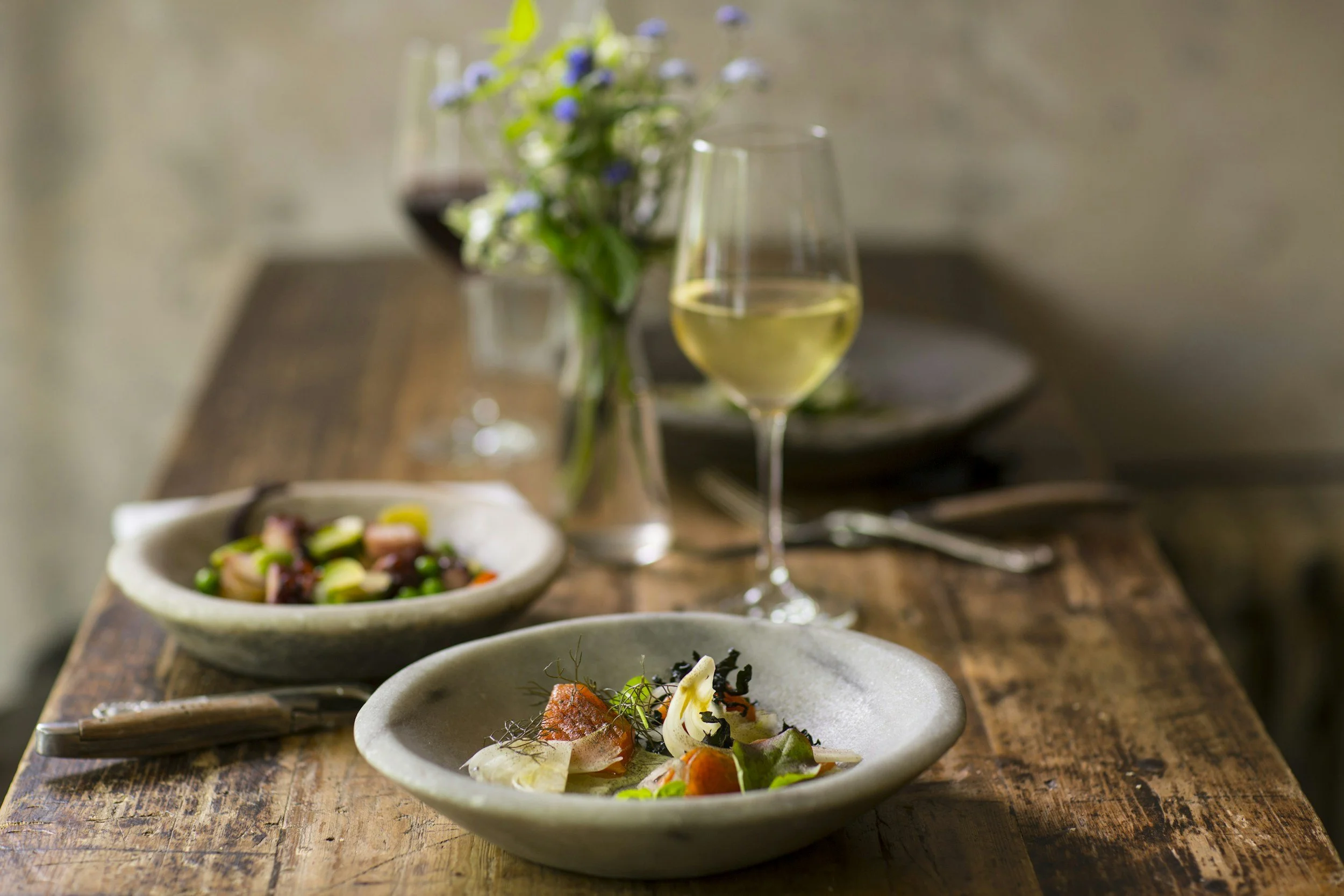 A rustic wooden table set with two bowls of food, a glass of white wine, a glass of red wine, and a small vase with blue and white flowers in the background.