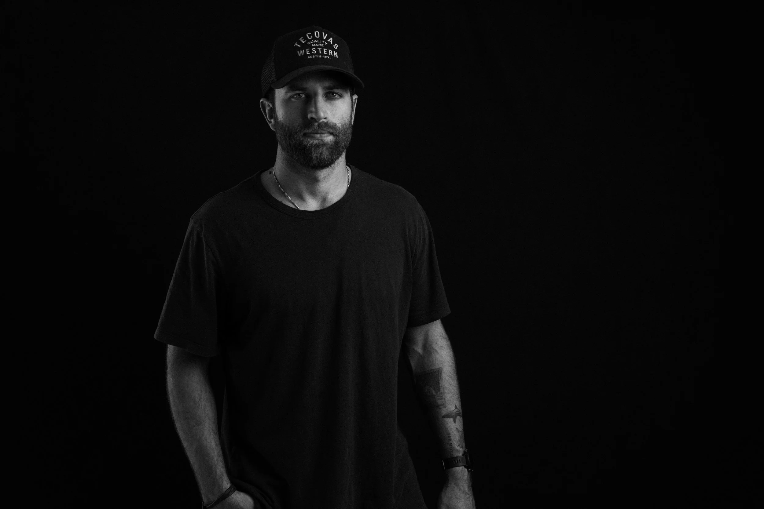 Black and white photo of a man with a beard wearing a black t-shirt and a baseball cap against a dark background.